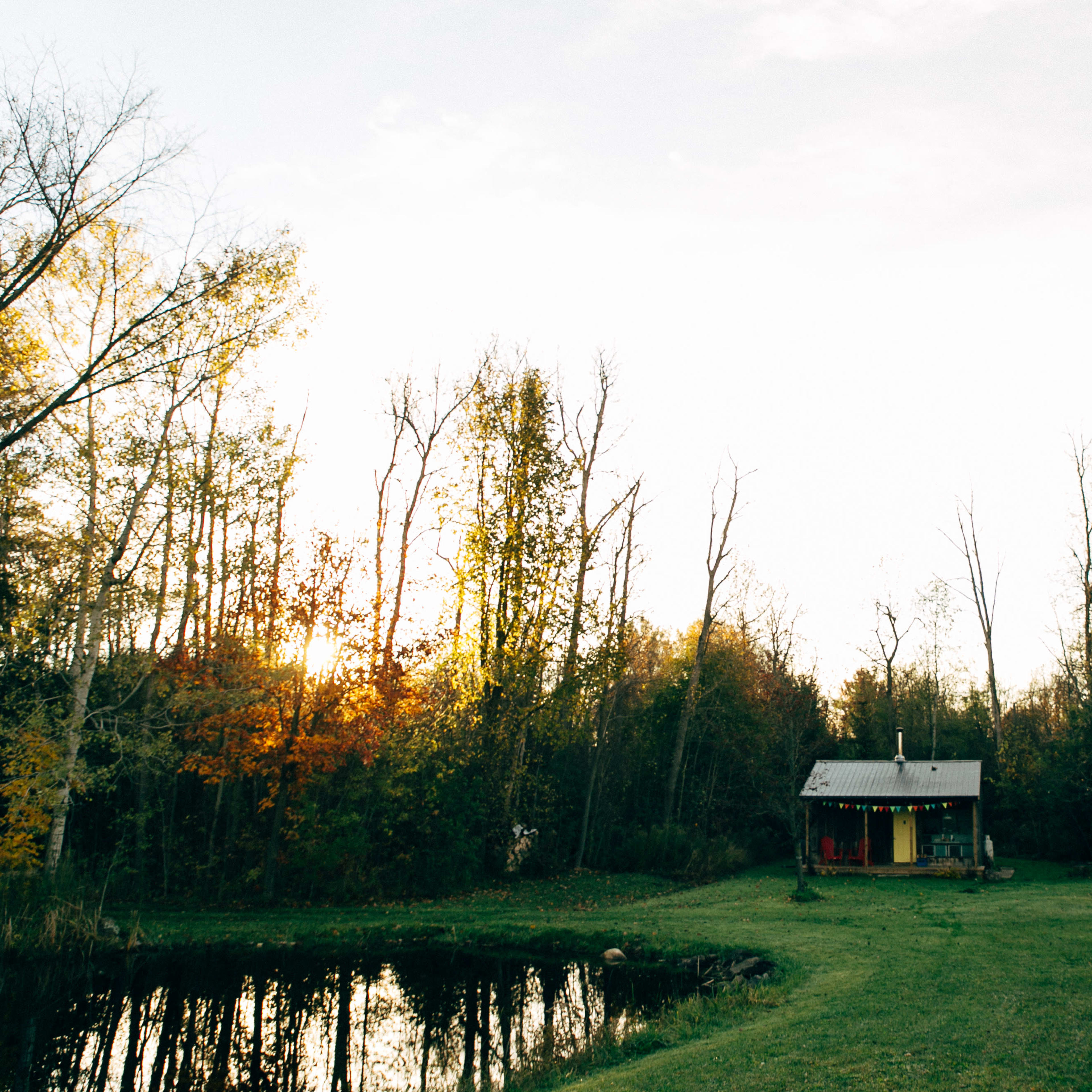 A small cabin with a metal roof is positioned near a pond, surrounded by trees as the sun sets in the background.