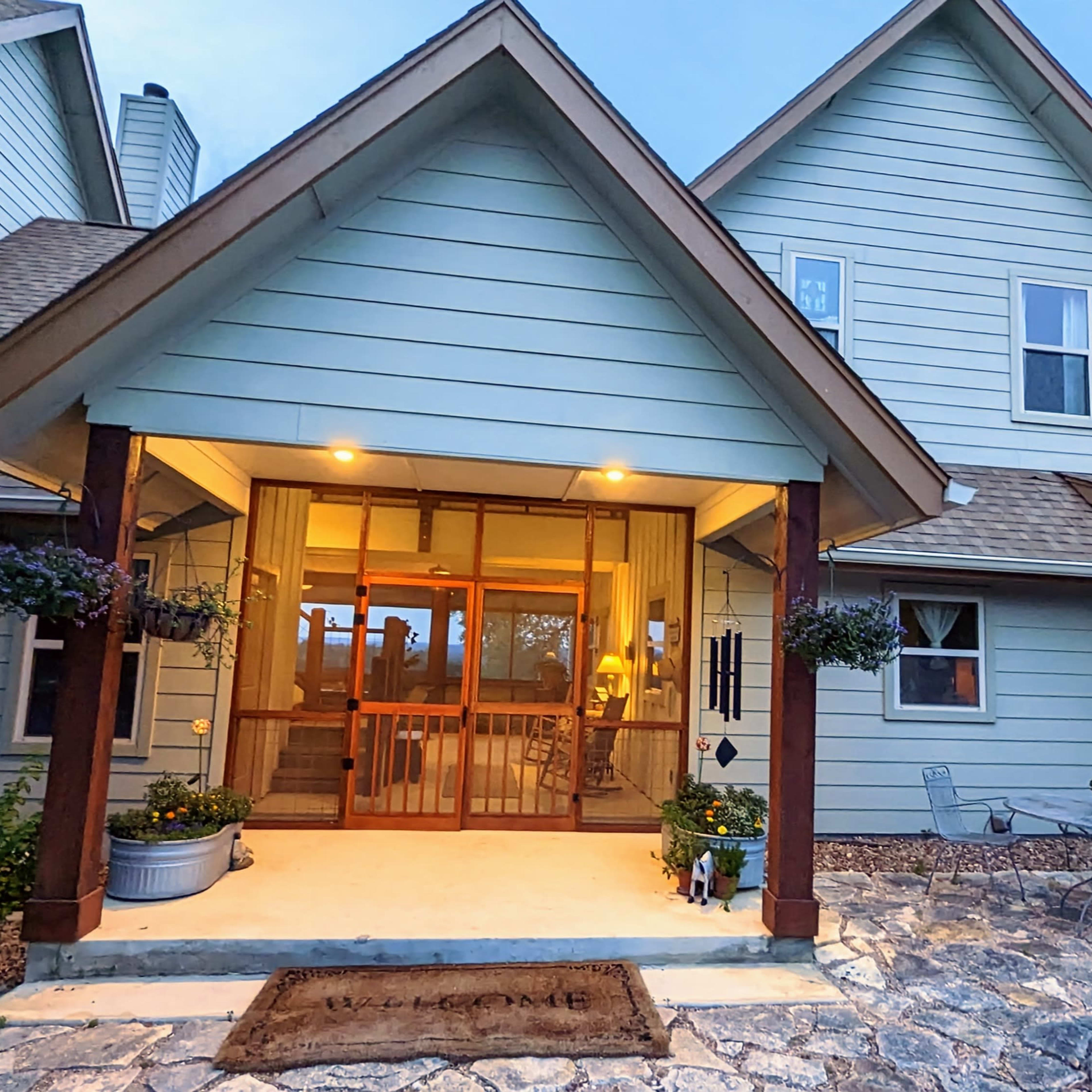 The entrance of a light blue house features a covered porch, stone walkway, and decorative potted plants.