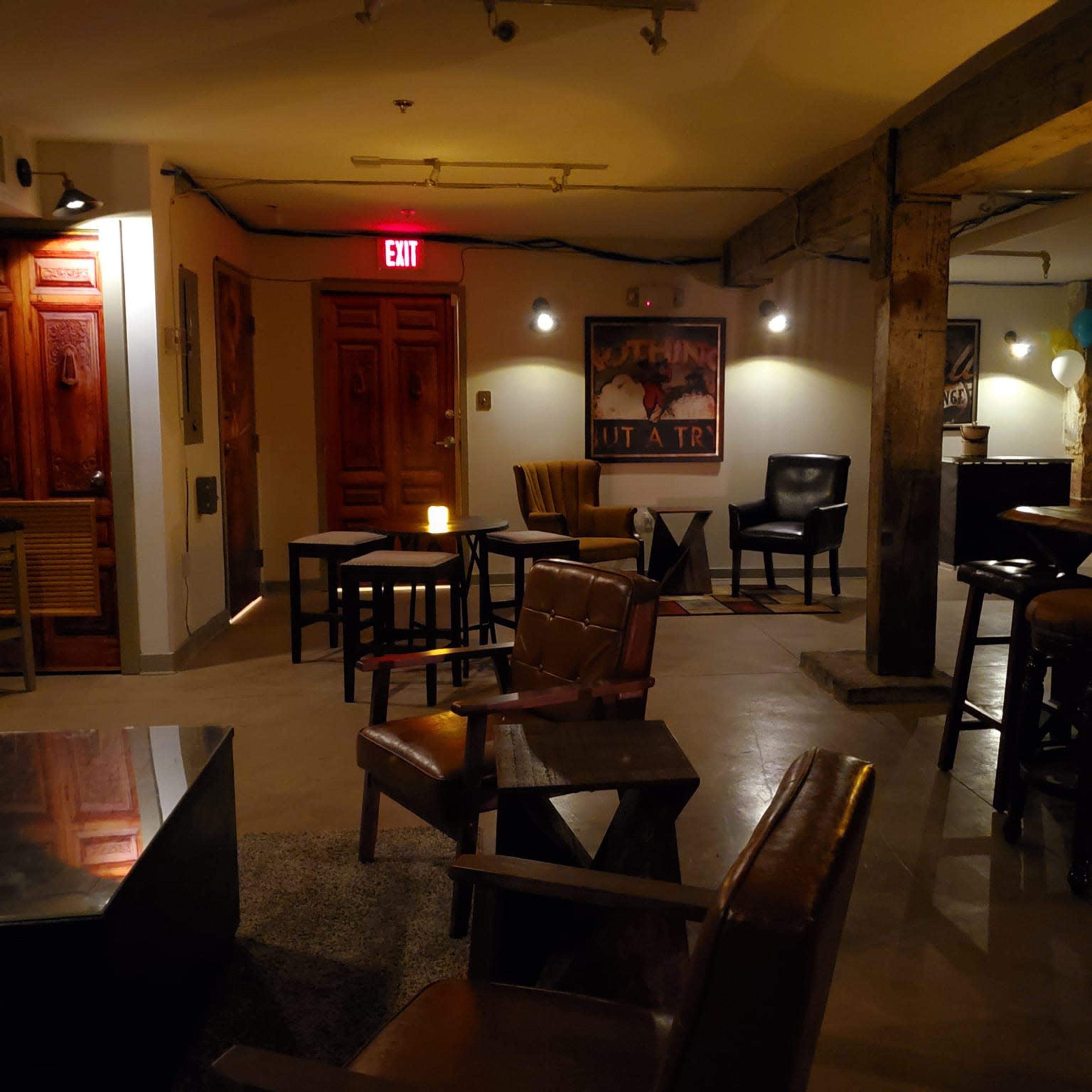 A dimly lit lounge area with various seating arrangements, including chairs and tables, and exposed brick walls.