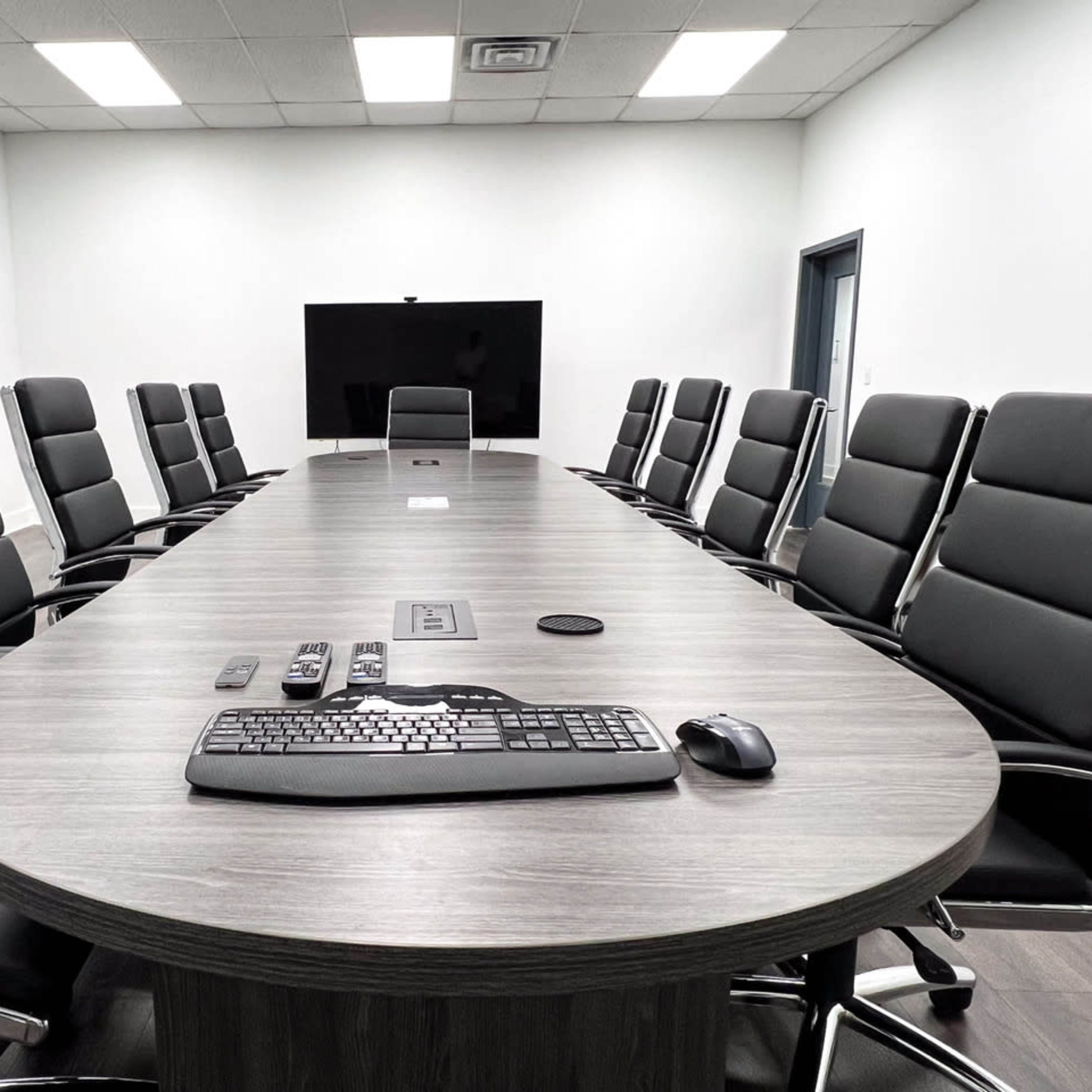 A long wooden conference table is surrounded by black leather chairs in a bright, minimalistic meeting room, featuring a large screen at one end.