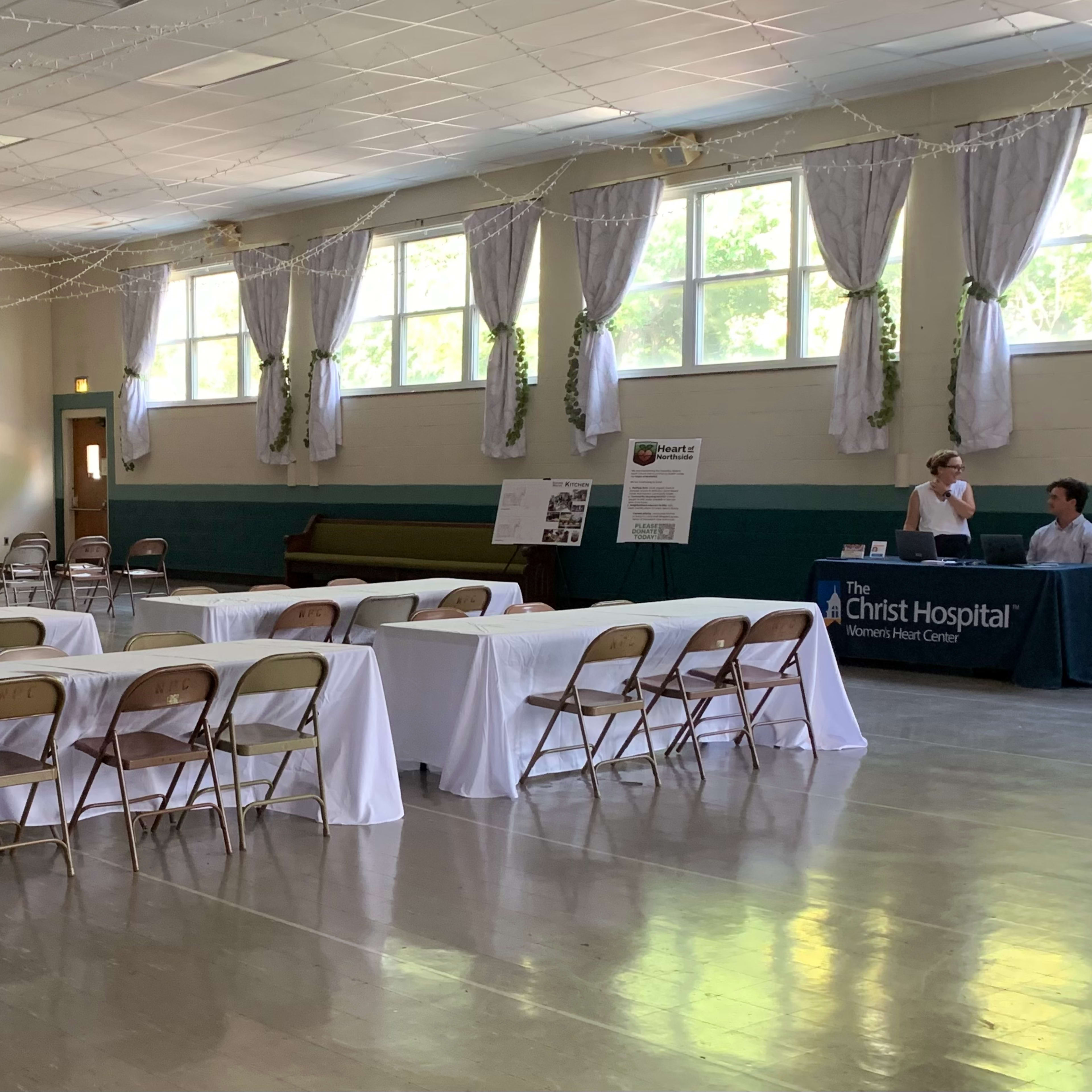 A spacious indoor venue features rows of chairs and tables, with a projector screen in the background and a booth set up in a corner.