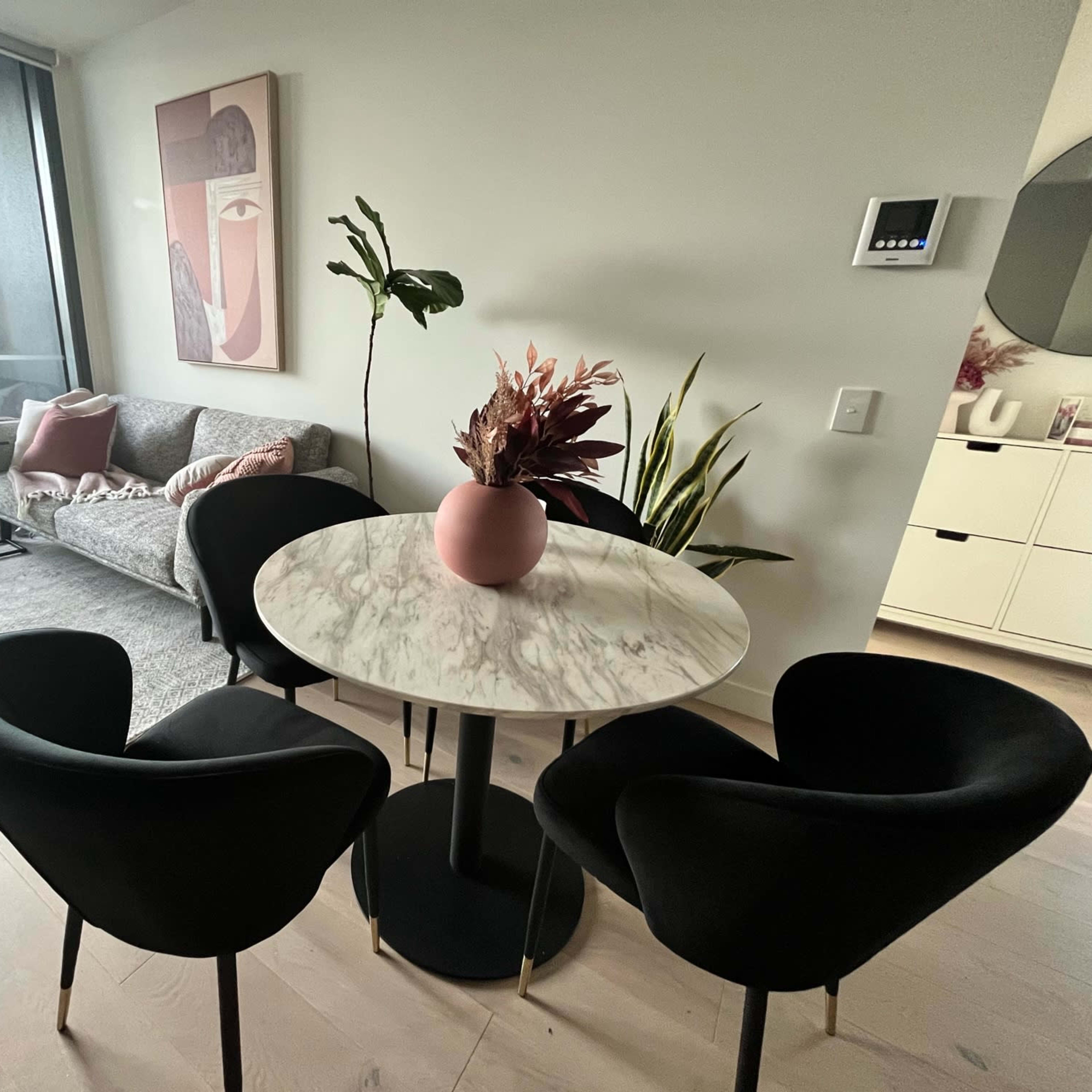 A modern living space featuring a marble dining table surrounded by four black chairs, with a gray sofa and decorative plants in the background.