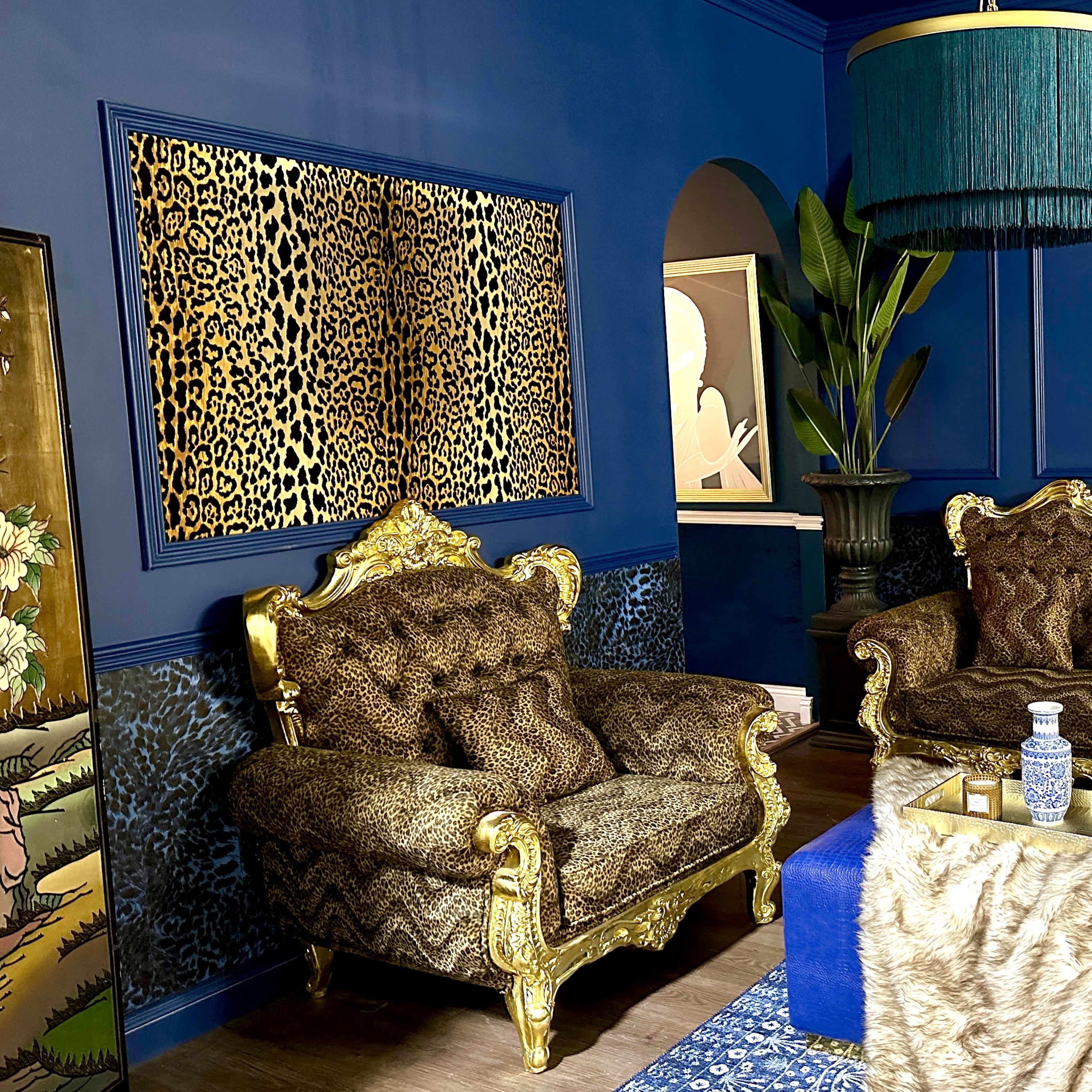 A room with dark blue walls, leopard print upholstery on two ornate armchairs, a large decorative plant, and abstract art on the wall.