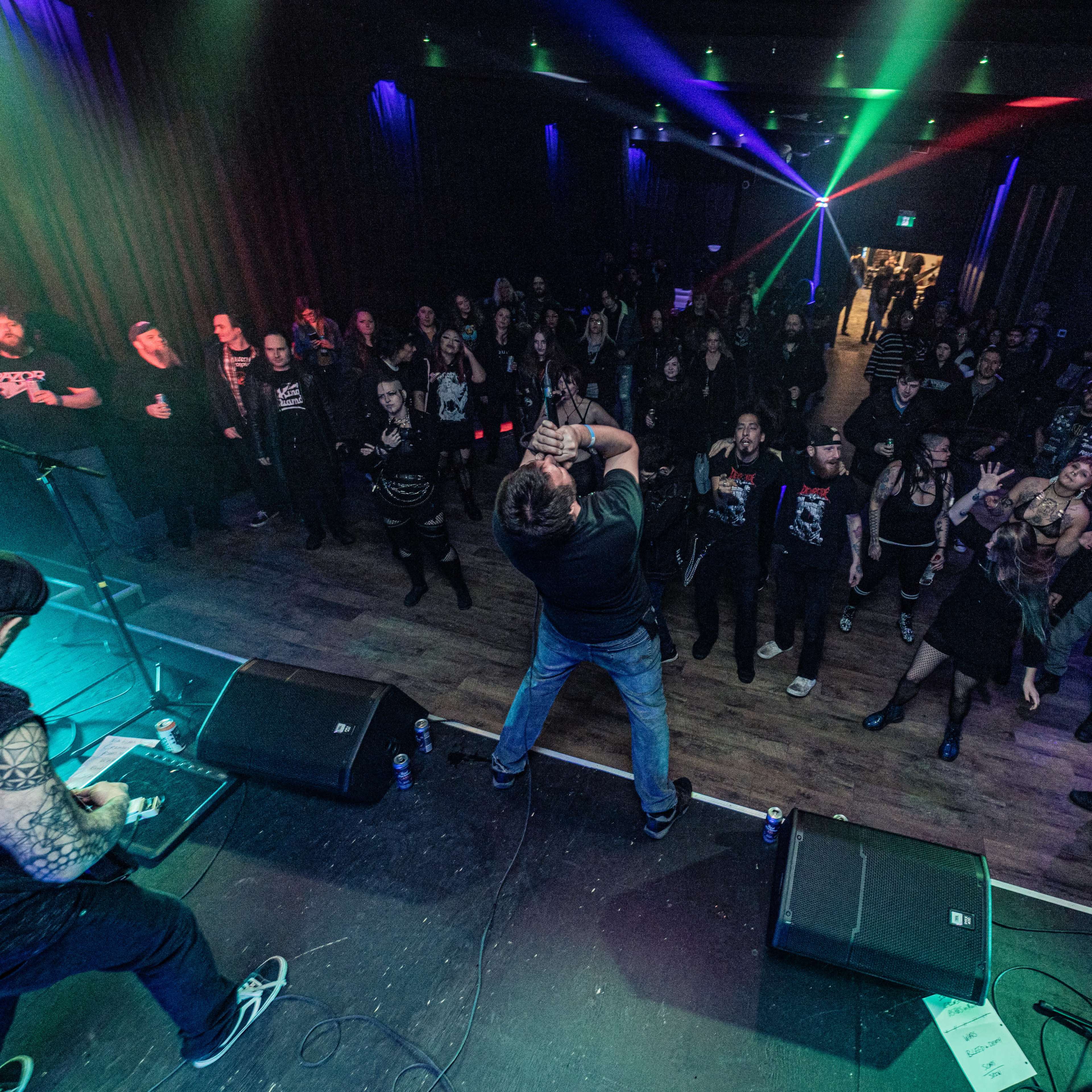 A live band performs on stage while an audience engages in front of them, illuminated by colorful laser lights in a dimly lit venue.