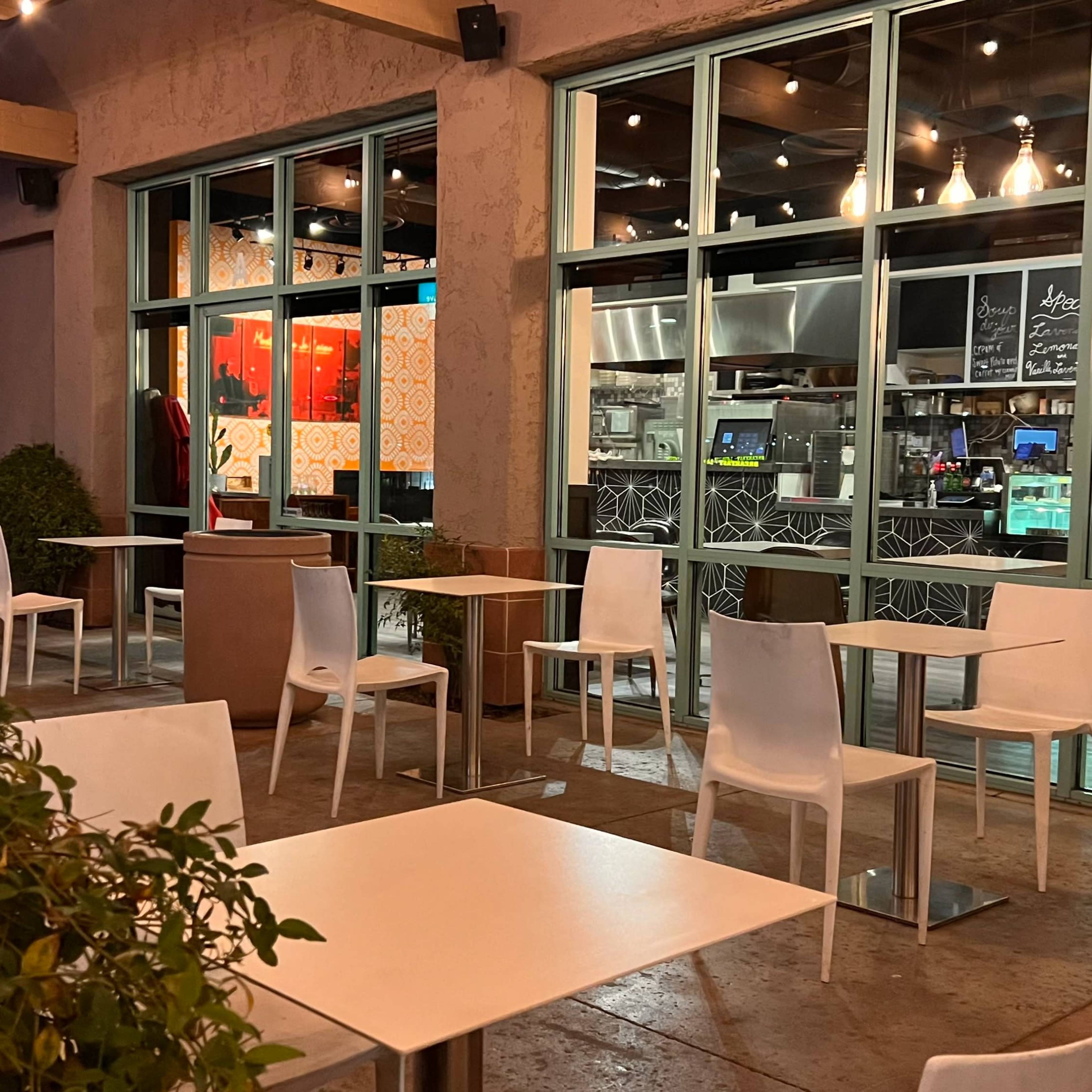 A partially outdoor dining area with empty white chairs and tables, overlooking a brightly lit restaurant interior.