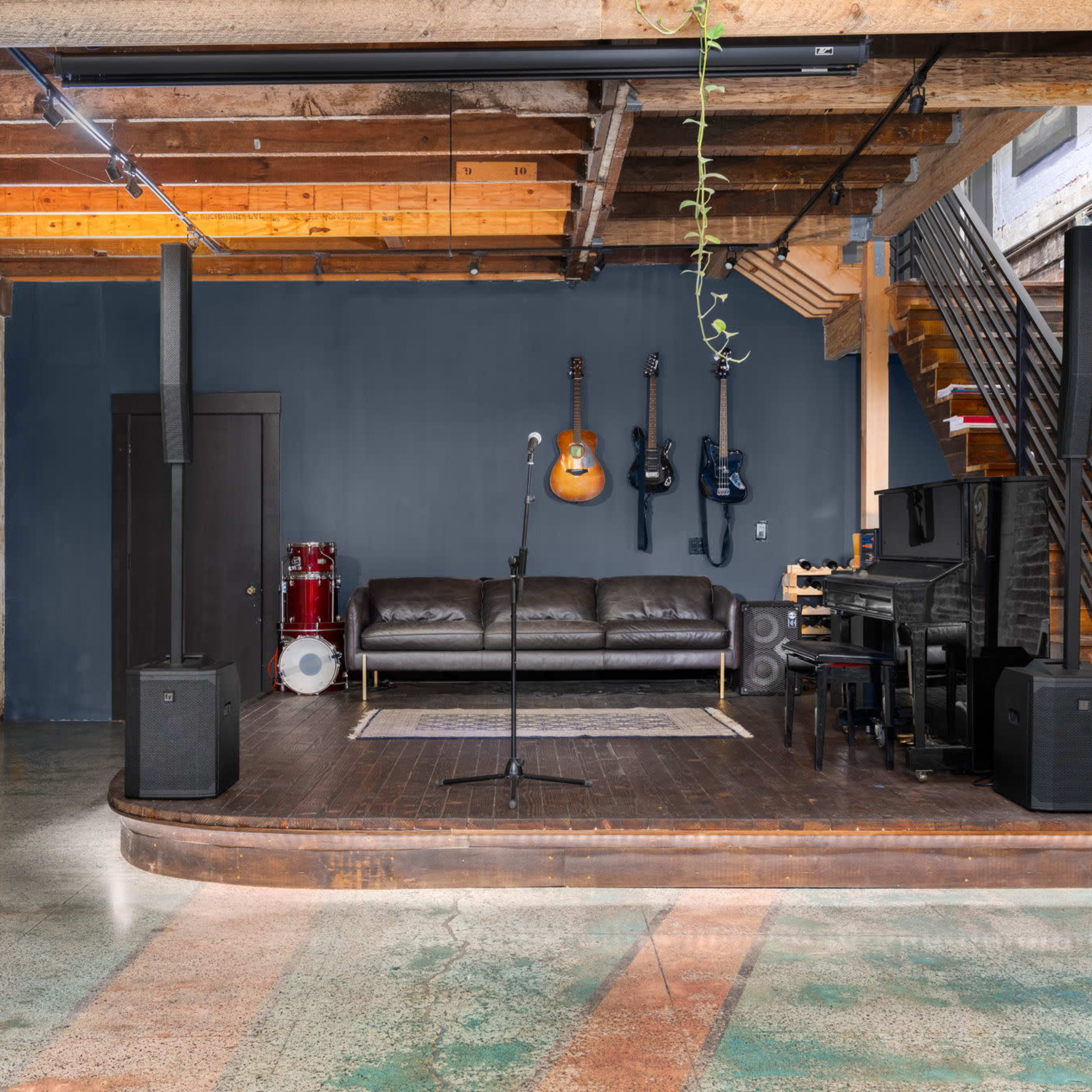 A spacious room with exposed brick walls, a wooden stage area featuring musical instruments, a black piano, and a staircase leading to an upper level.
