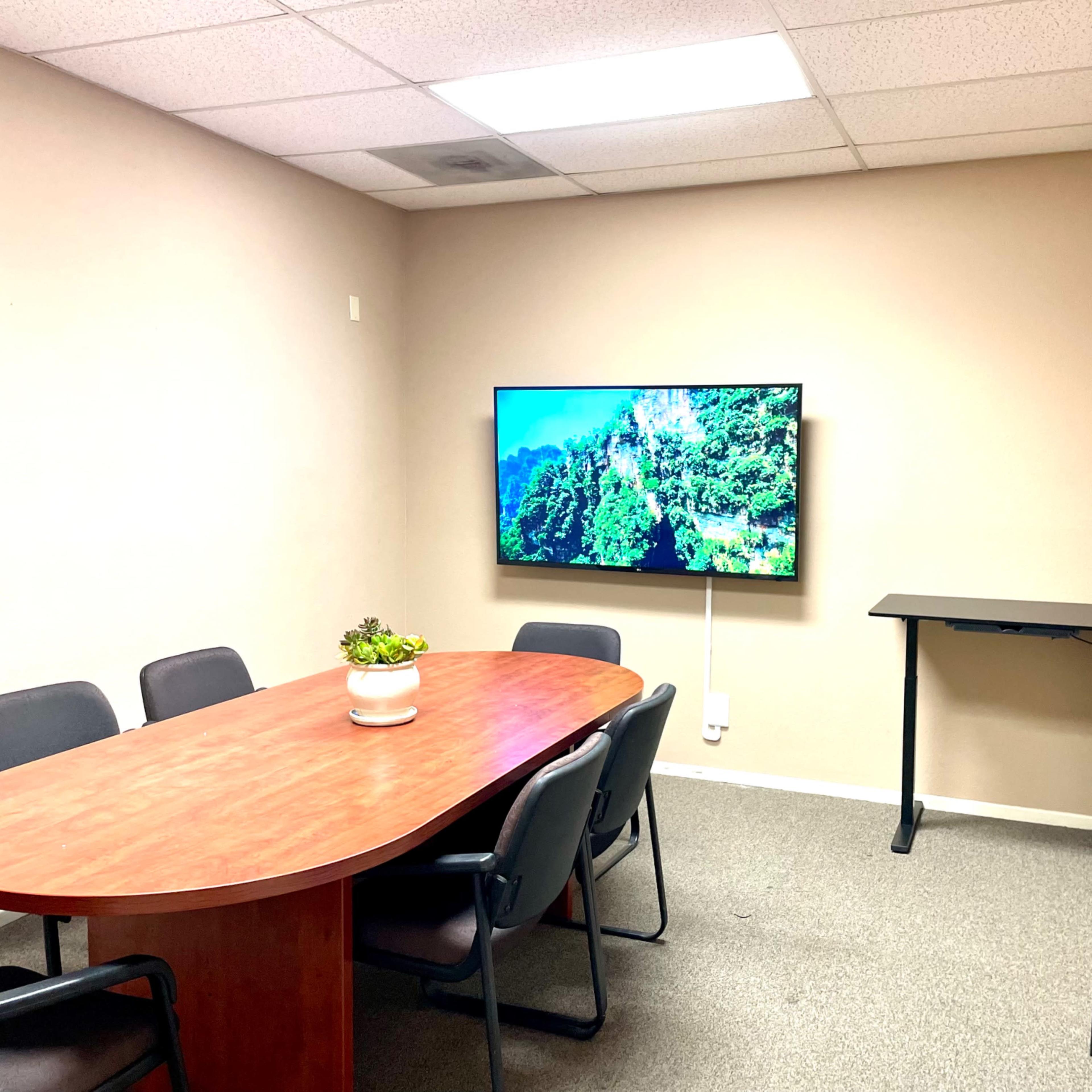 A small conference room with a wooden oval table, six black chairs, a mounted television displaying a nature scene, and a standing desk against one wall.
