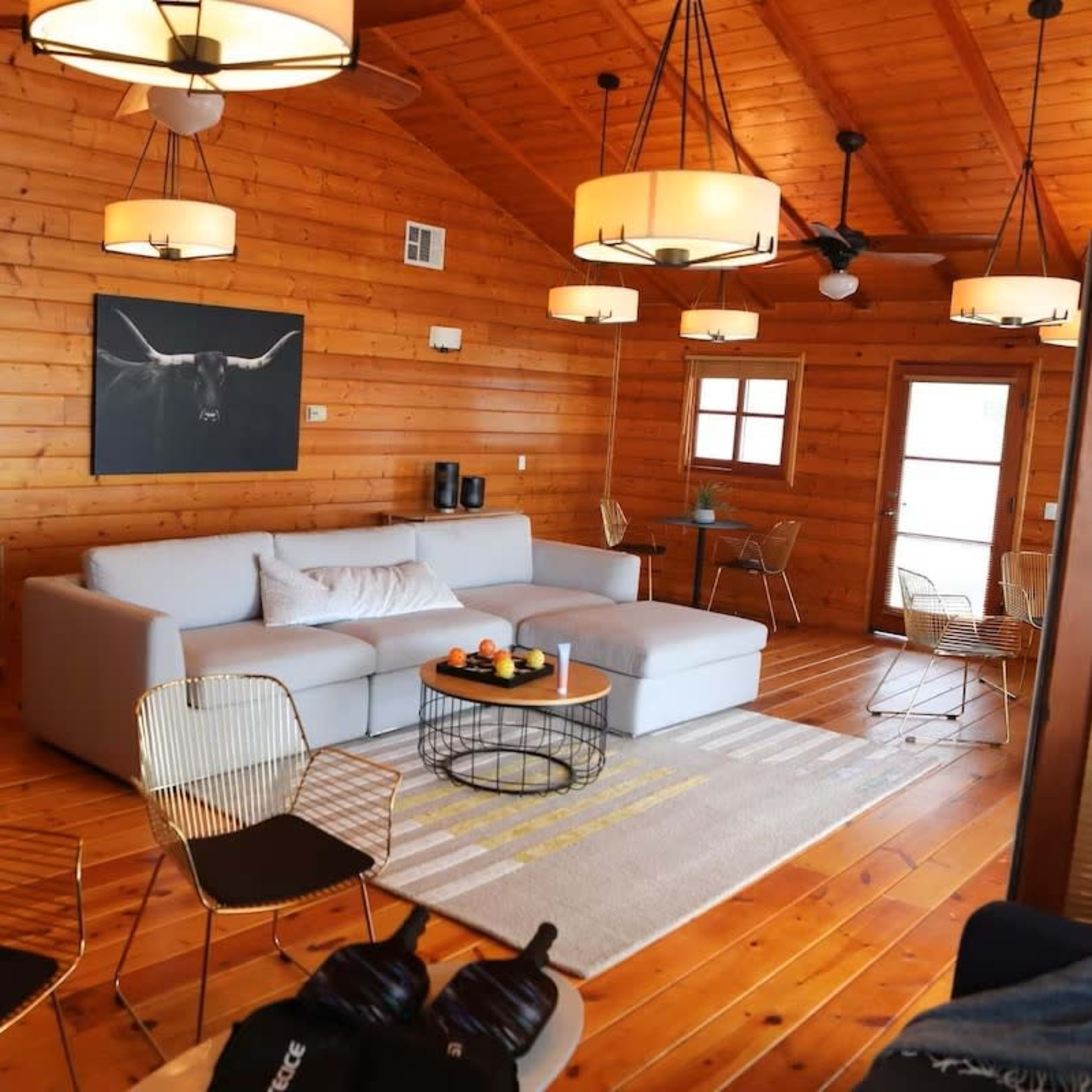 A cozy wooden cabin living room with a large gray sectional sofa, a coffee table with snacks, and metal chairs arranged around a light-colored rug.