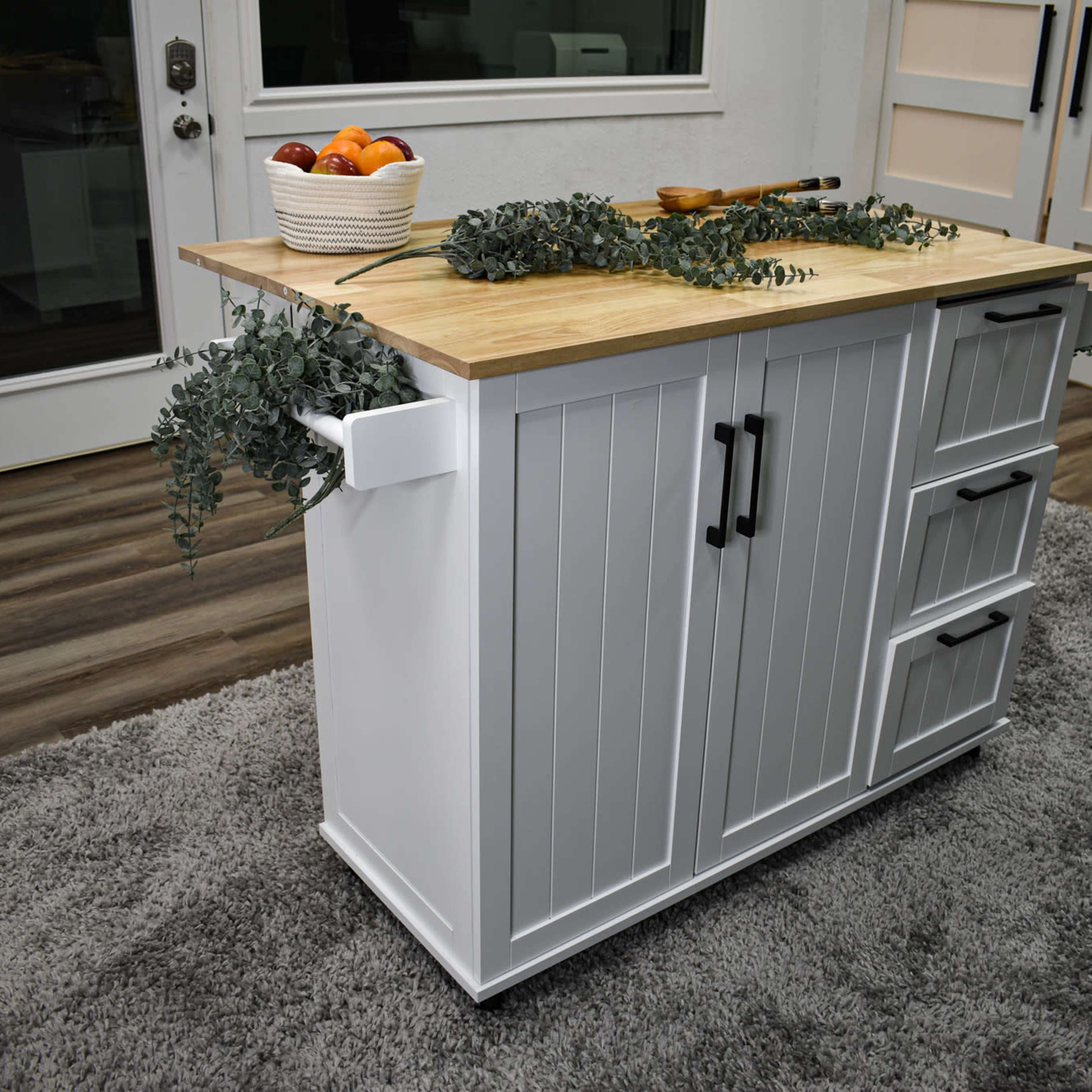 A white kitchen island with a wooden countertop is adorned with a bowl of fruit and greenery, positioned on a gray carpeted floor.