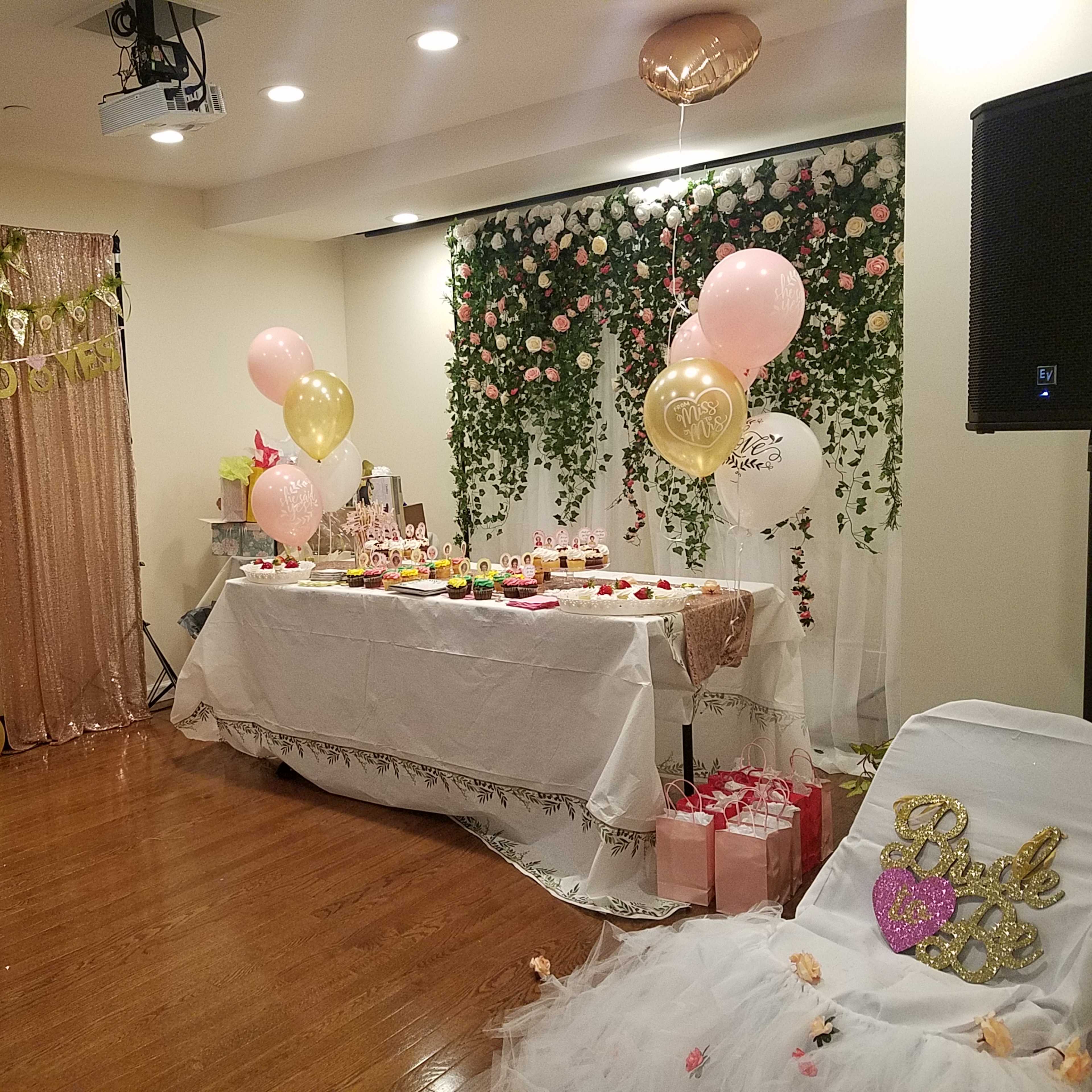 The scene features a decorated event space with a table laden with treats, a backdrop of pink and gold decorations, and balloons amidst a floral wall.