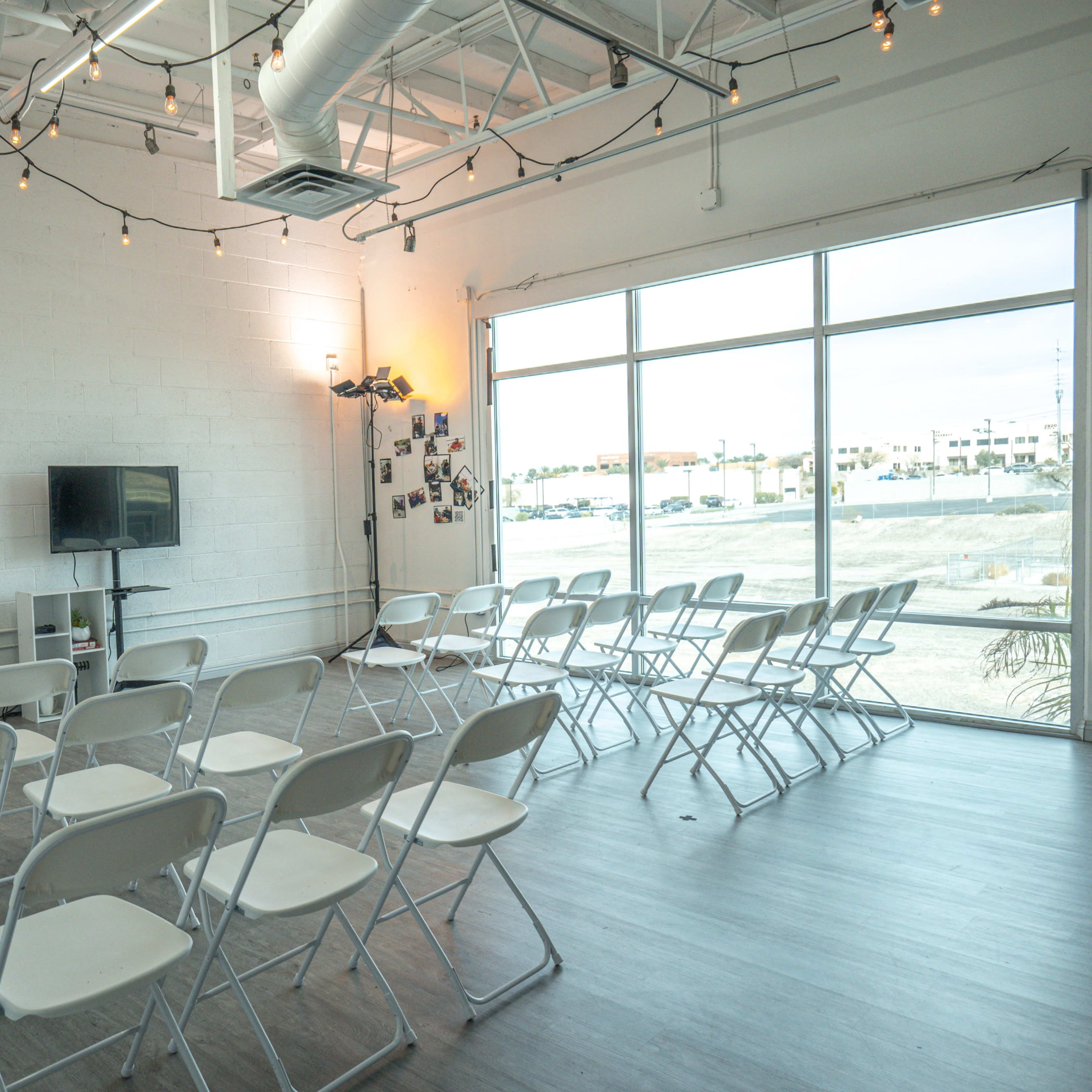 A spacious room arranged with white folding chairs facing a television mounted on a wall, with a large window and string lights overhead.
