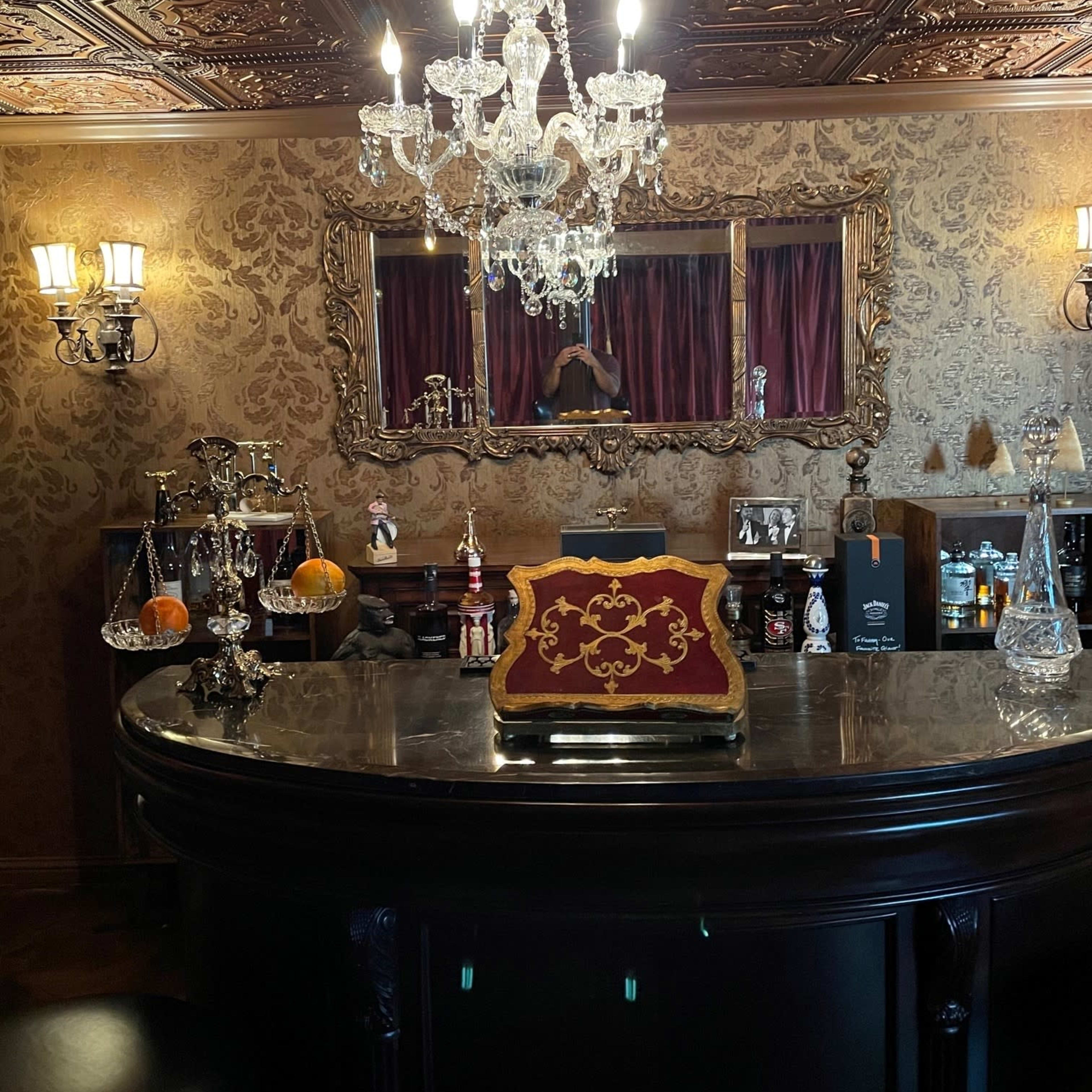 A well-appointed home bar with a chandelier, ornate wall decor, and an elegantly styled counter displaying various bottles and glassware.