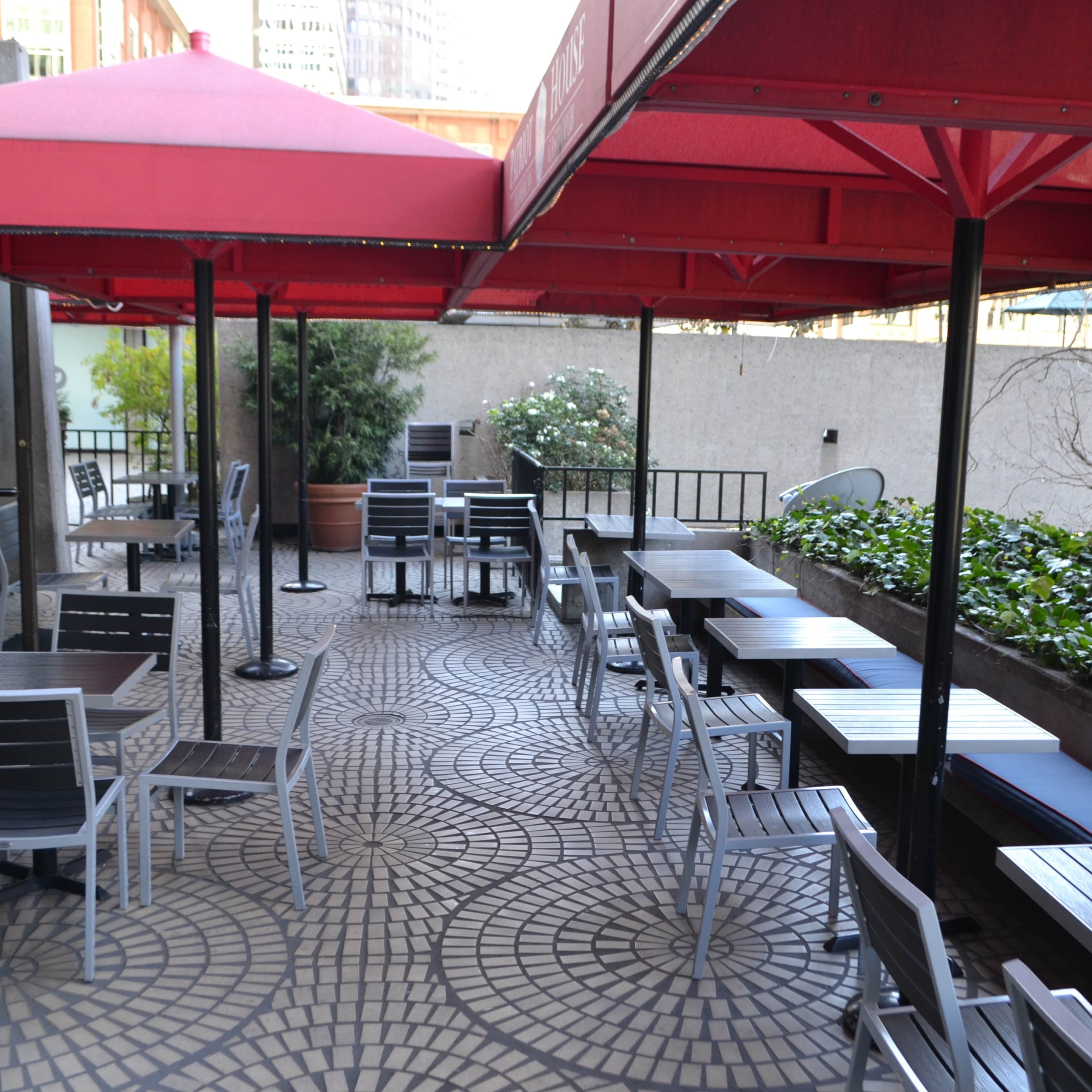 An outdoor seating area with red umbrellas, featuring gray and white tables and chairs arranged on a patterned tile surface.