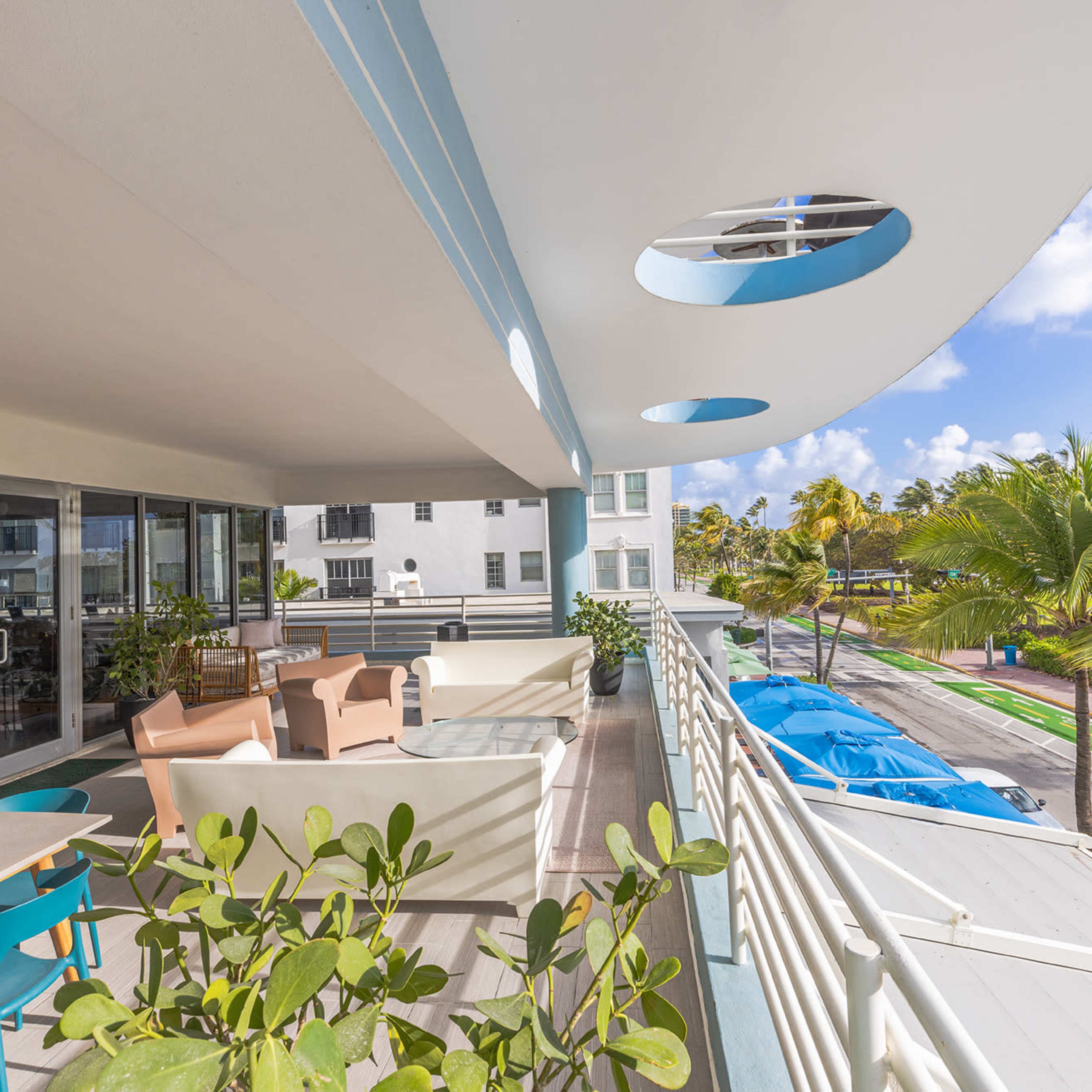 A modern balcony with outdoor furniture, potted plants, and a view of a palm-lined street and parked boats.