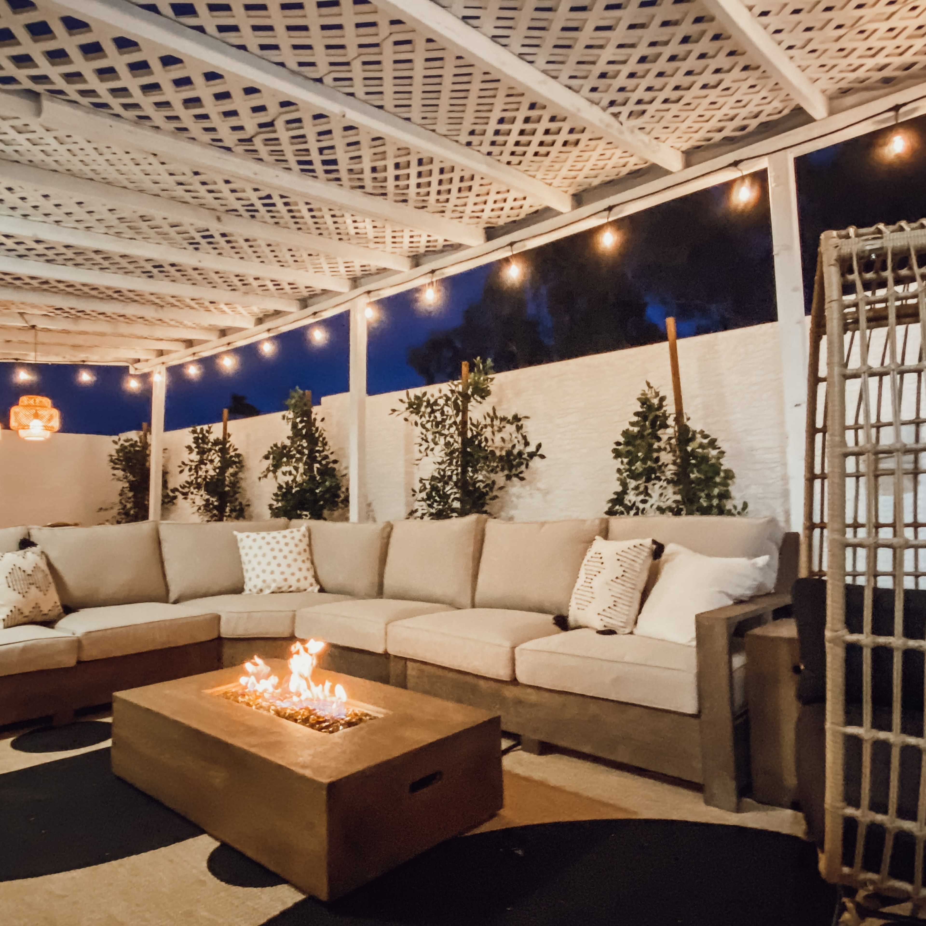 A spacious outdoor patio area features a large sectional sofa surrounding a fire pit, with decorative string lights overhead and lush greenery nearby.