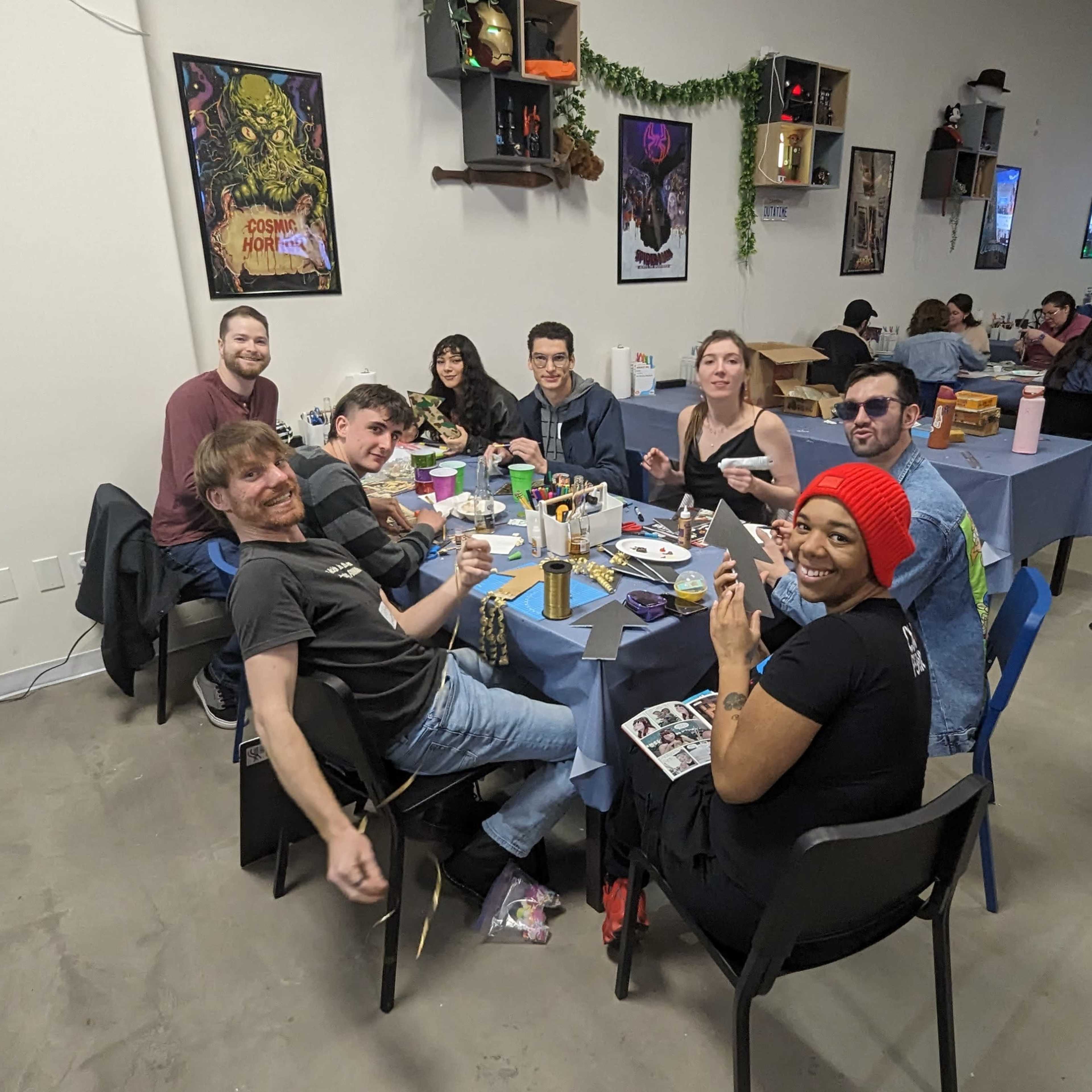 A group of nine people sits around a table filled with food and drinks in a decorated room with artwork and a TV in the background.