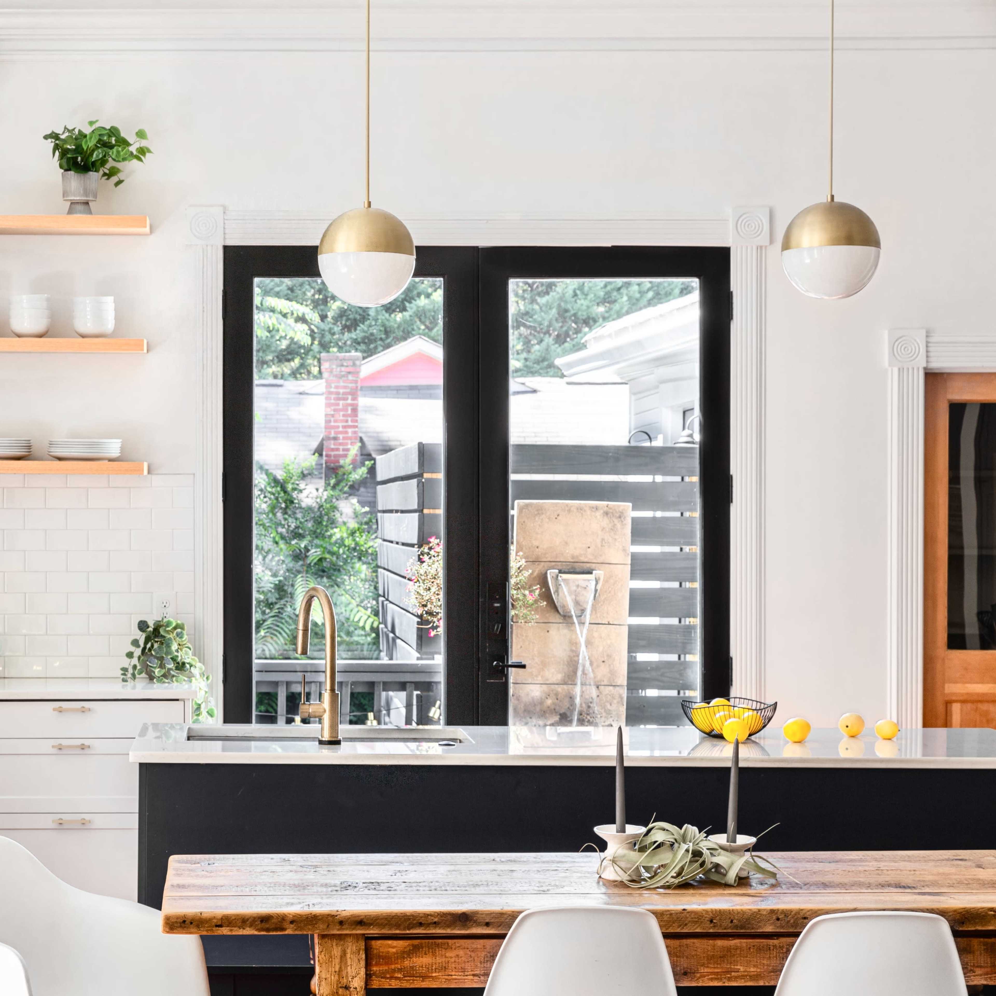 A modern kitchen features open shelving with dishware, a central wooden dining table, and large windows providing natural light.
