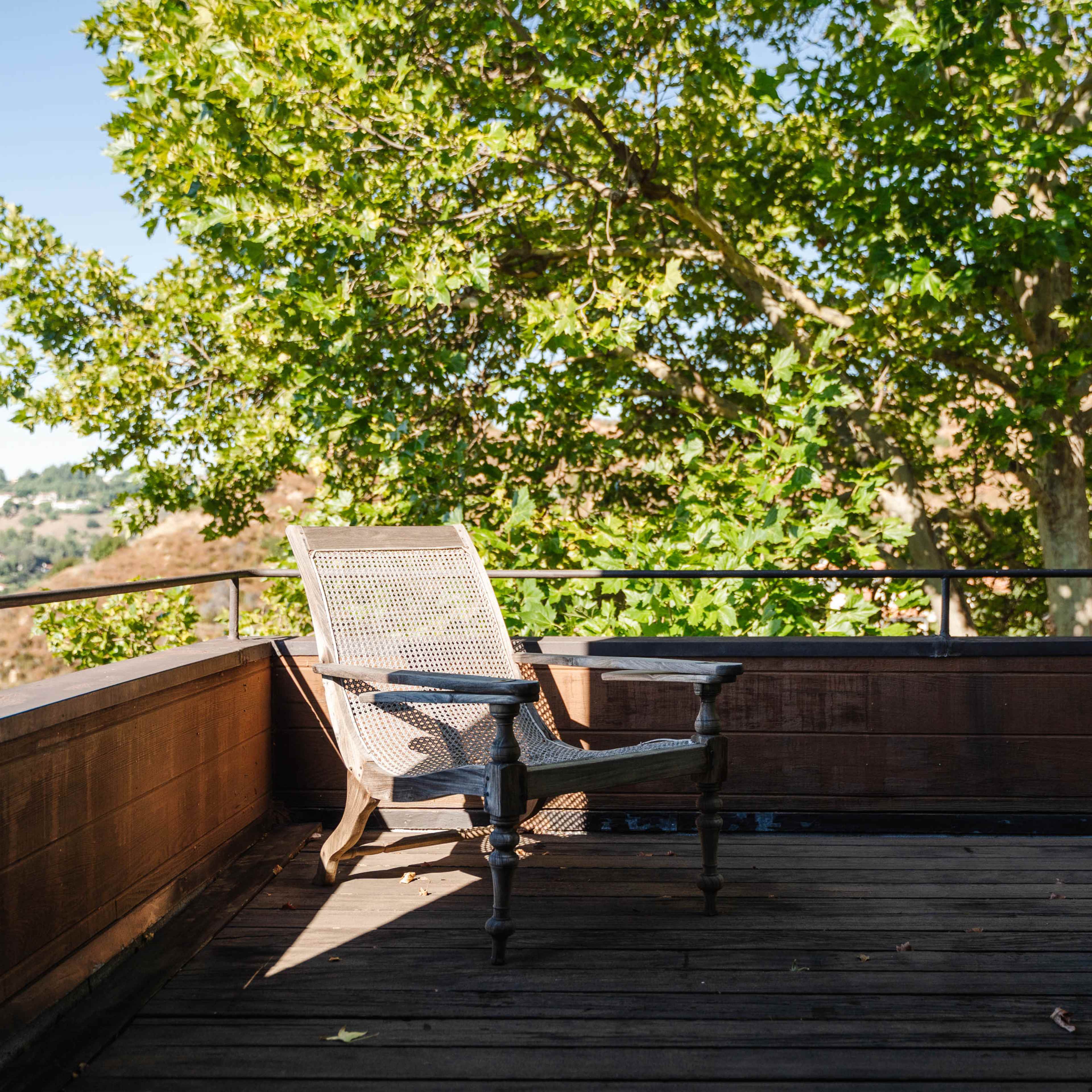 A wooden chair sits on a deck with a railing, surrounded by a tree and hills in the background.
