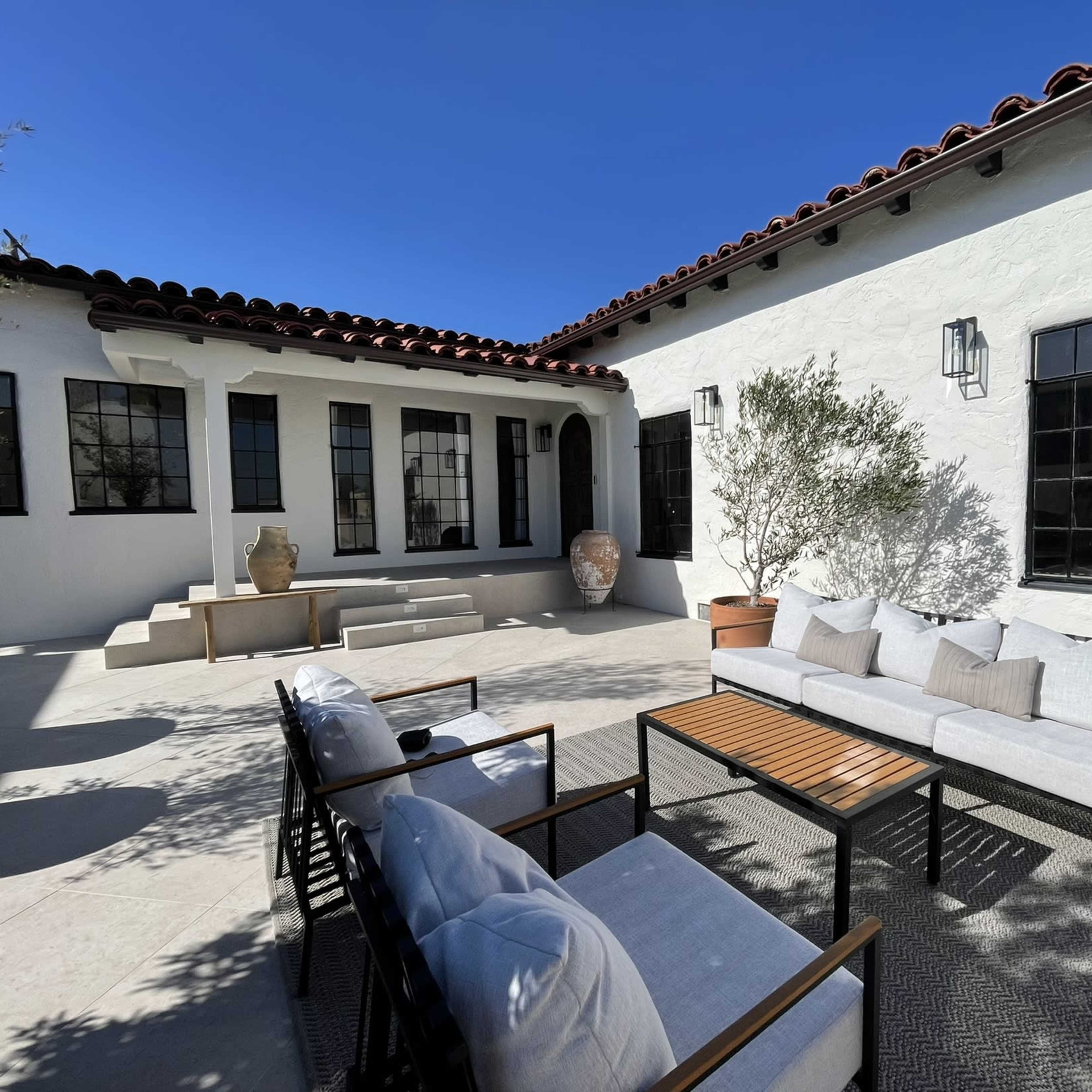 A patio with modern seating and a table is surrounded by white walls and large windows in a sunny courtyard.