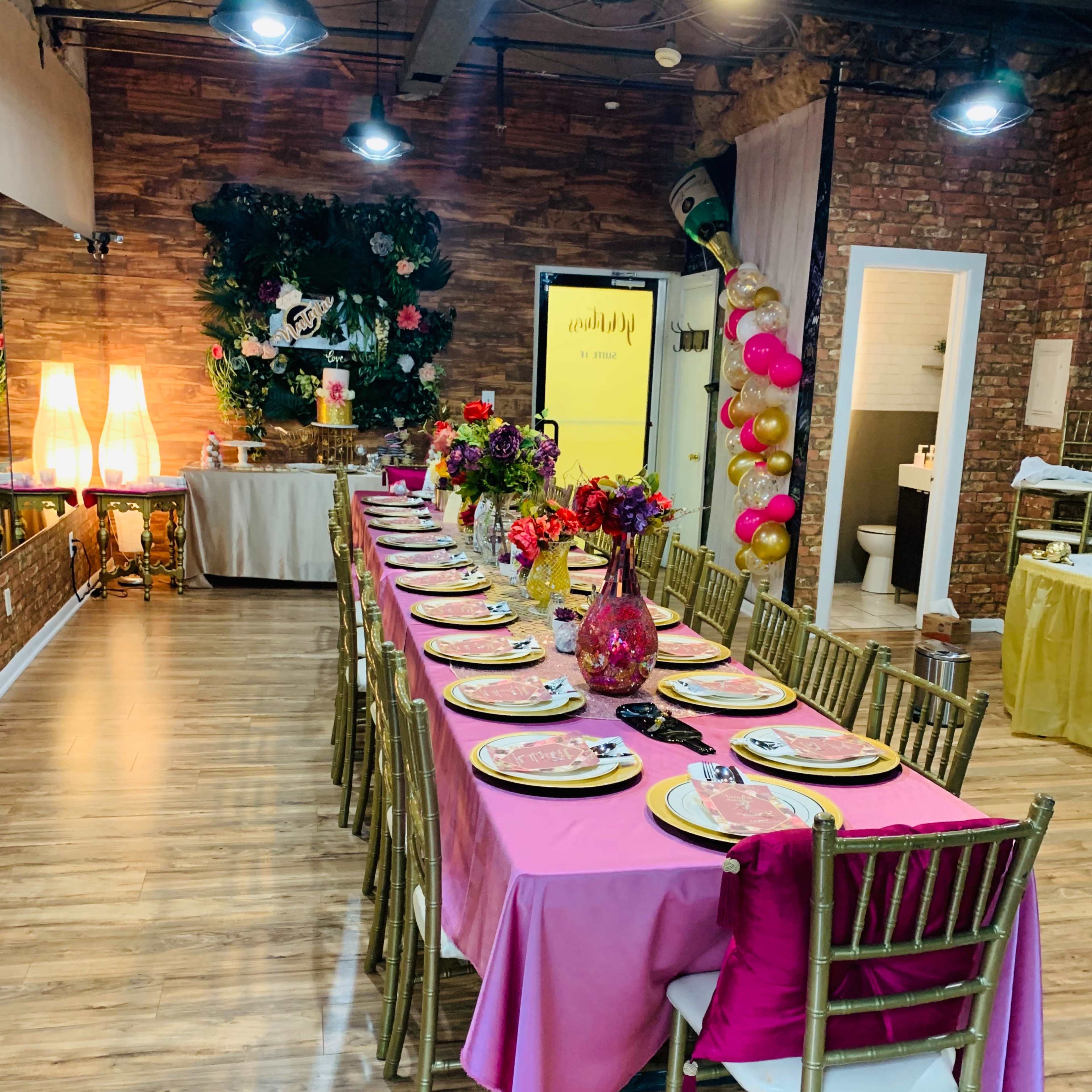 A long dining table is set with pink and gold tableware in a well-decorated event space featuring floral arrangements and a backdrop with balloons.