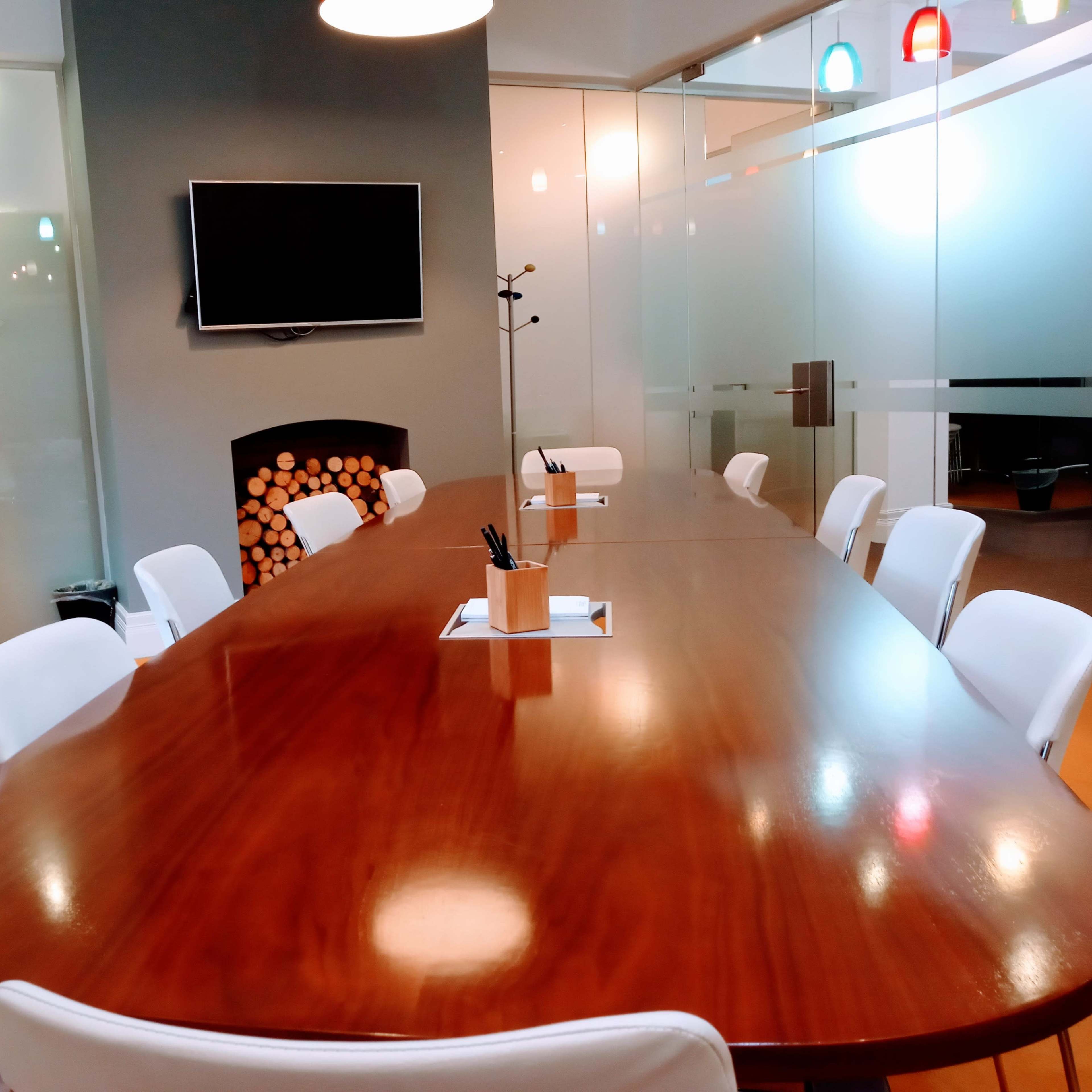 A modern conference room featuring a long wooden table, white chairs, a television on the wall, and a stack of logs in a decorative fireplace.