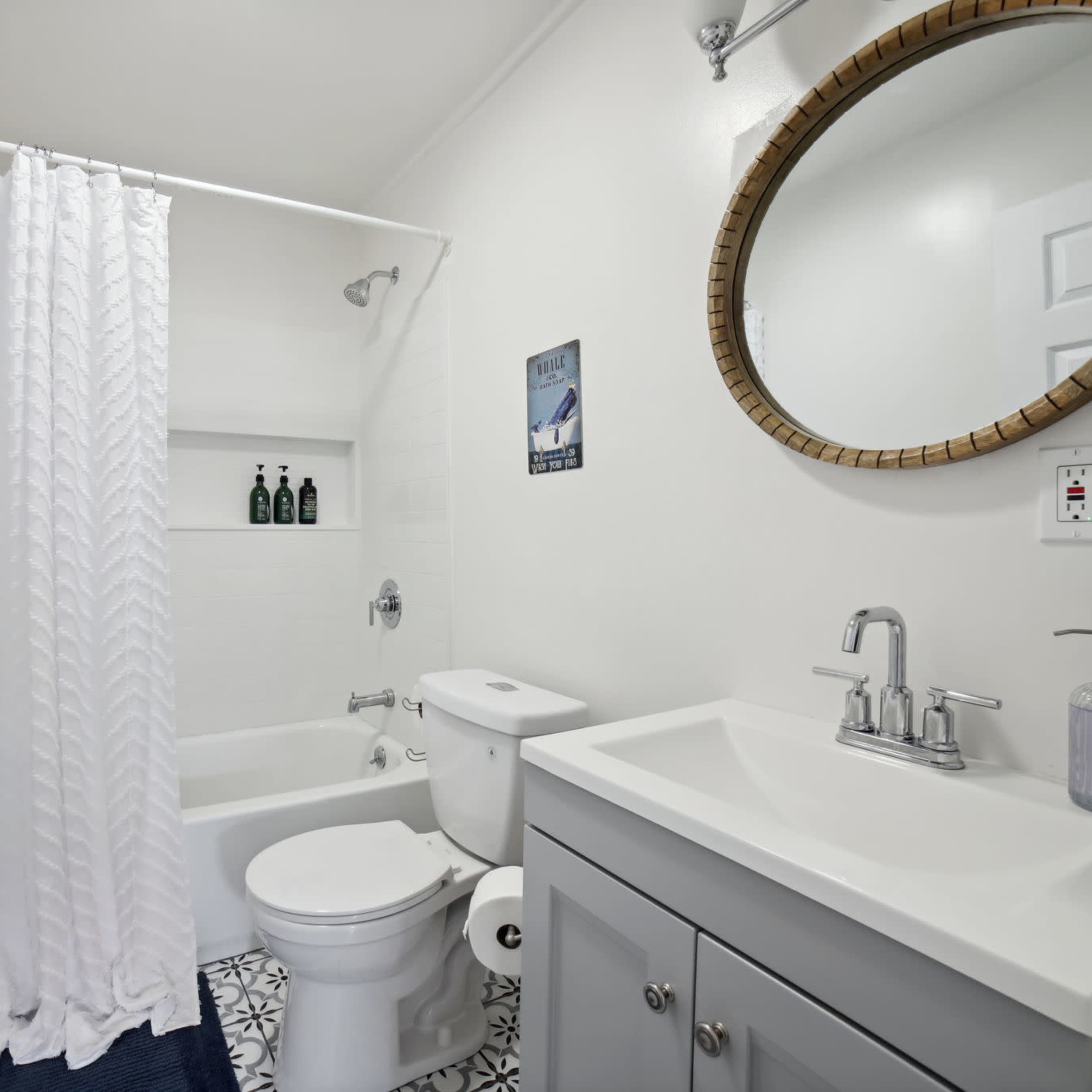 This bathroom features a bathtub with a shower curtain, a round mirror above a sink, and a toilet next to a small shelf displaying bottles.