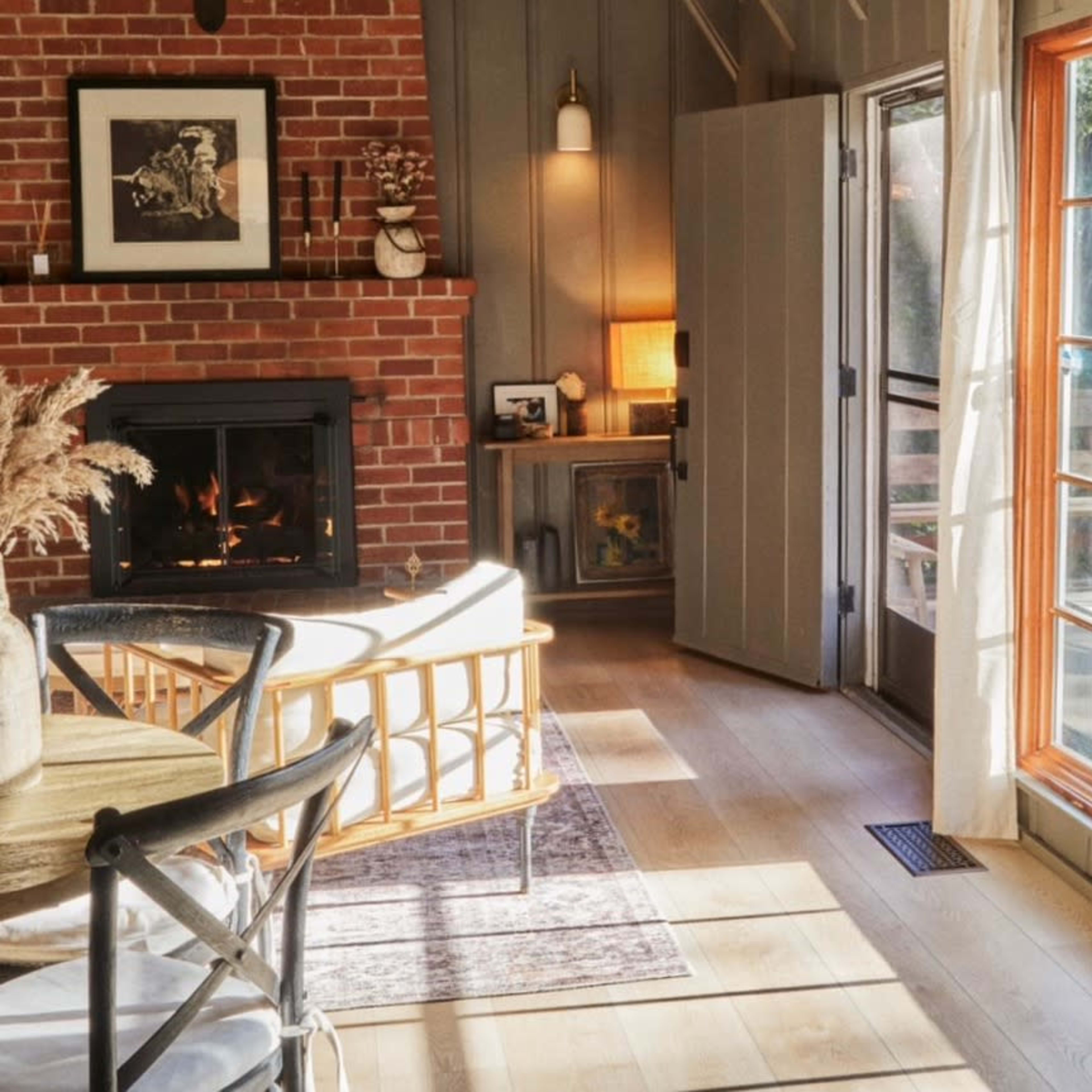 A cozy living area with a brick fireplace, a rattan chair, and large windows allowing natural light to flood the space.