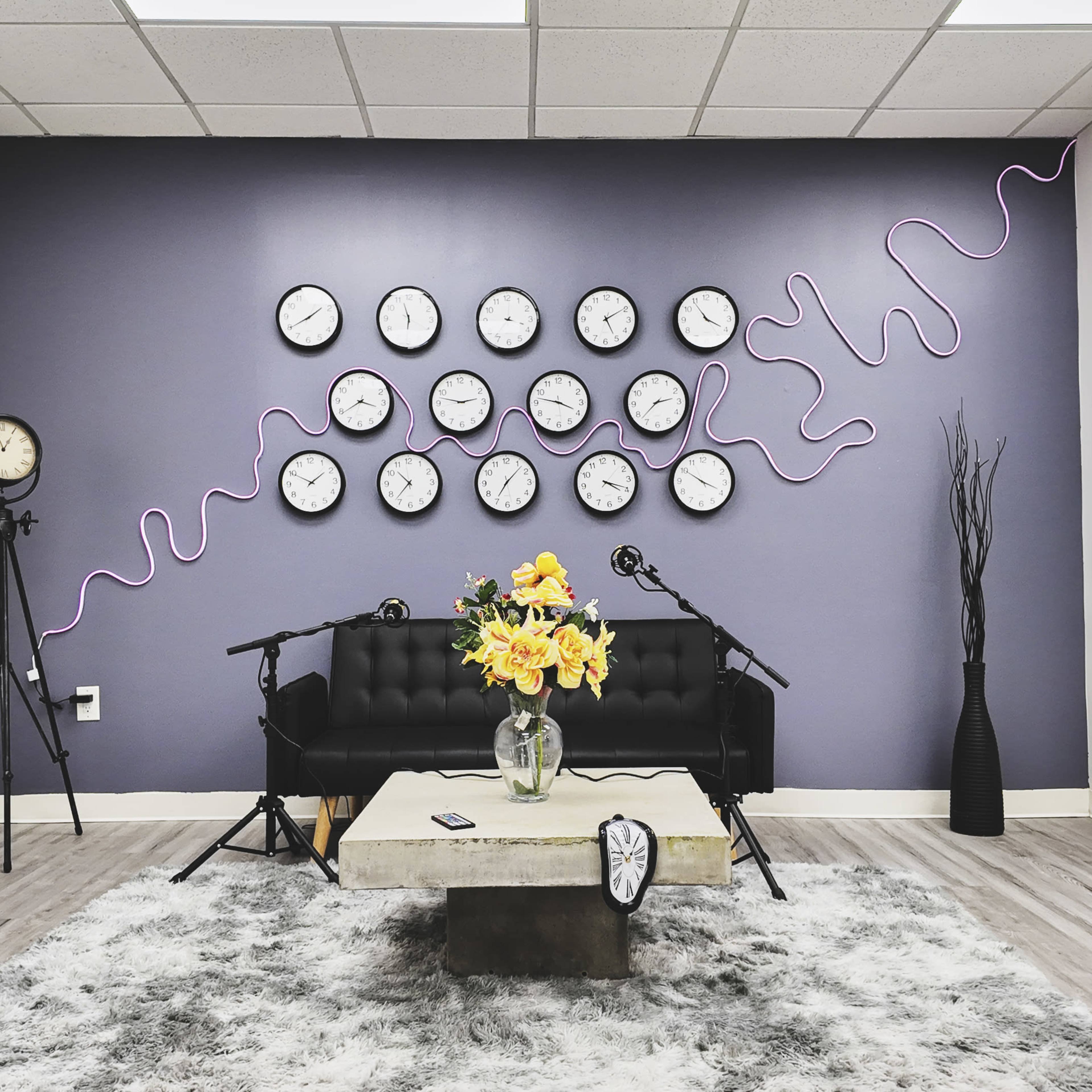 The room features a black couch with a coffee table and a large arrangement of yellow flowers, surrounded by multiple wall clocks on a purple backdrop and accent decor in a modern style.