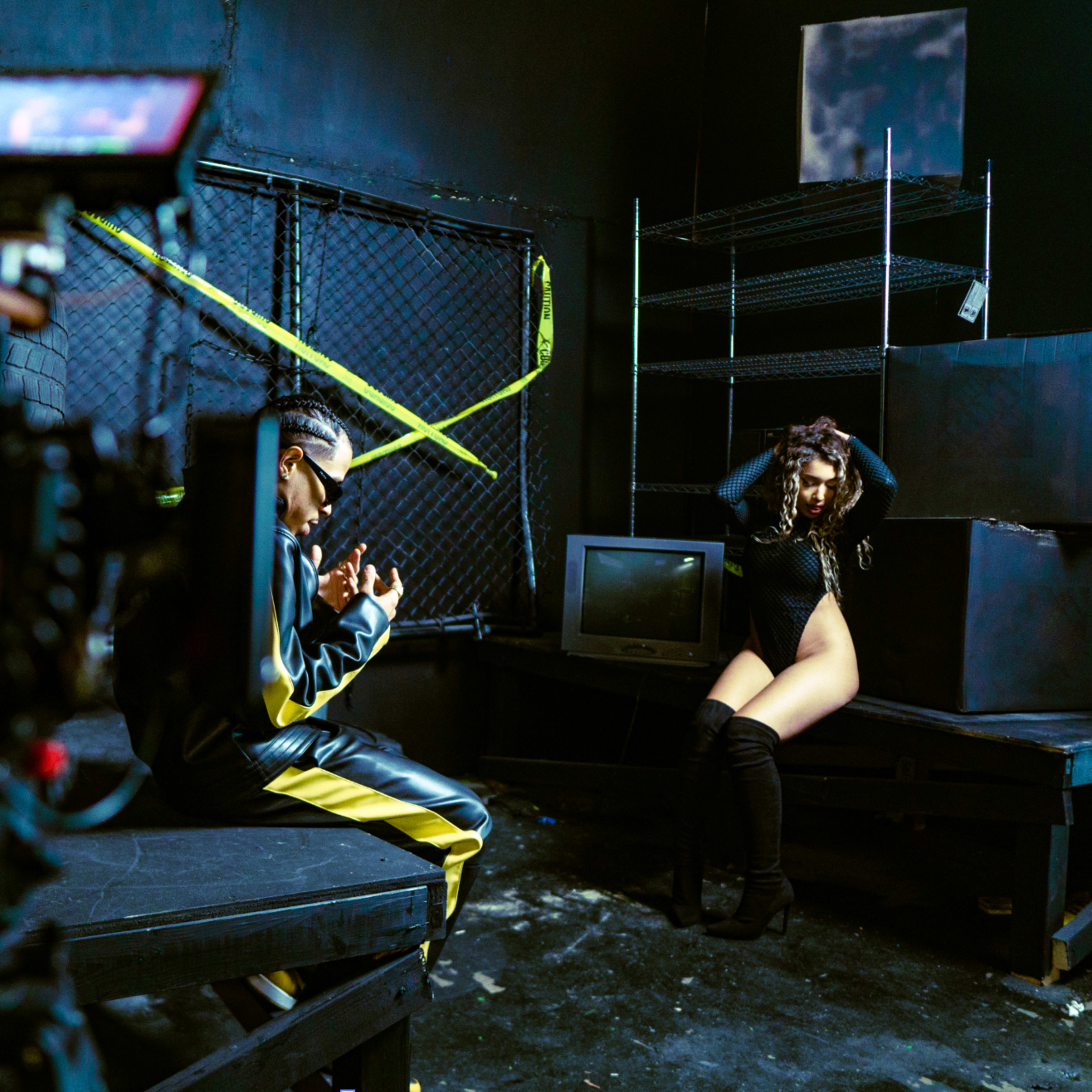 A photographer captures a model posing in a black outfit amidst tires and dark furniture in an enclosed space.