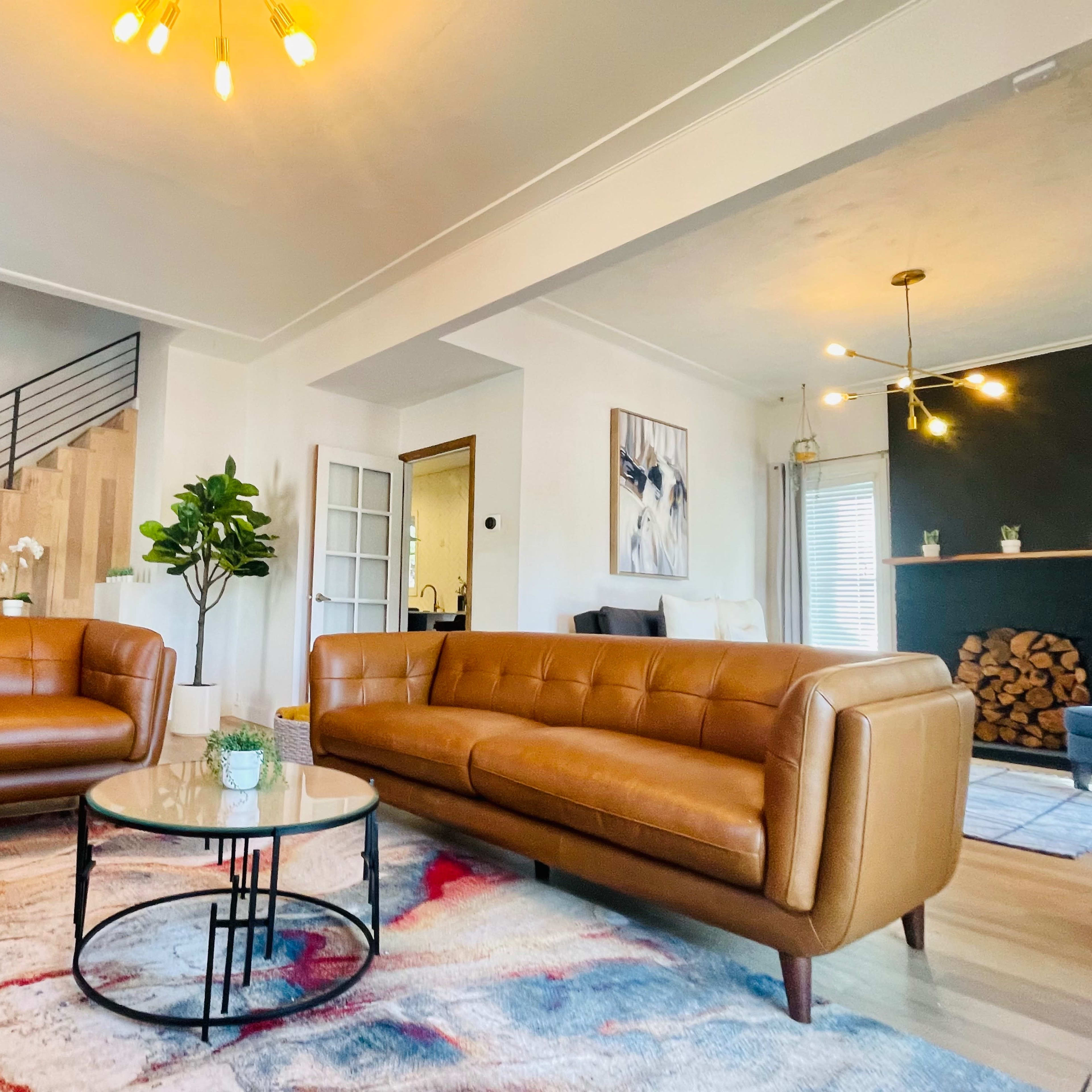 A living room features a brown leather sofa and loveseat around a circular glass coffee table on a patterned rug, with a staircase in the background.