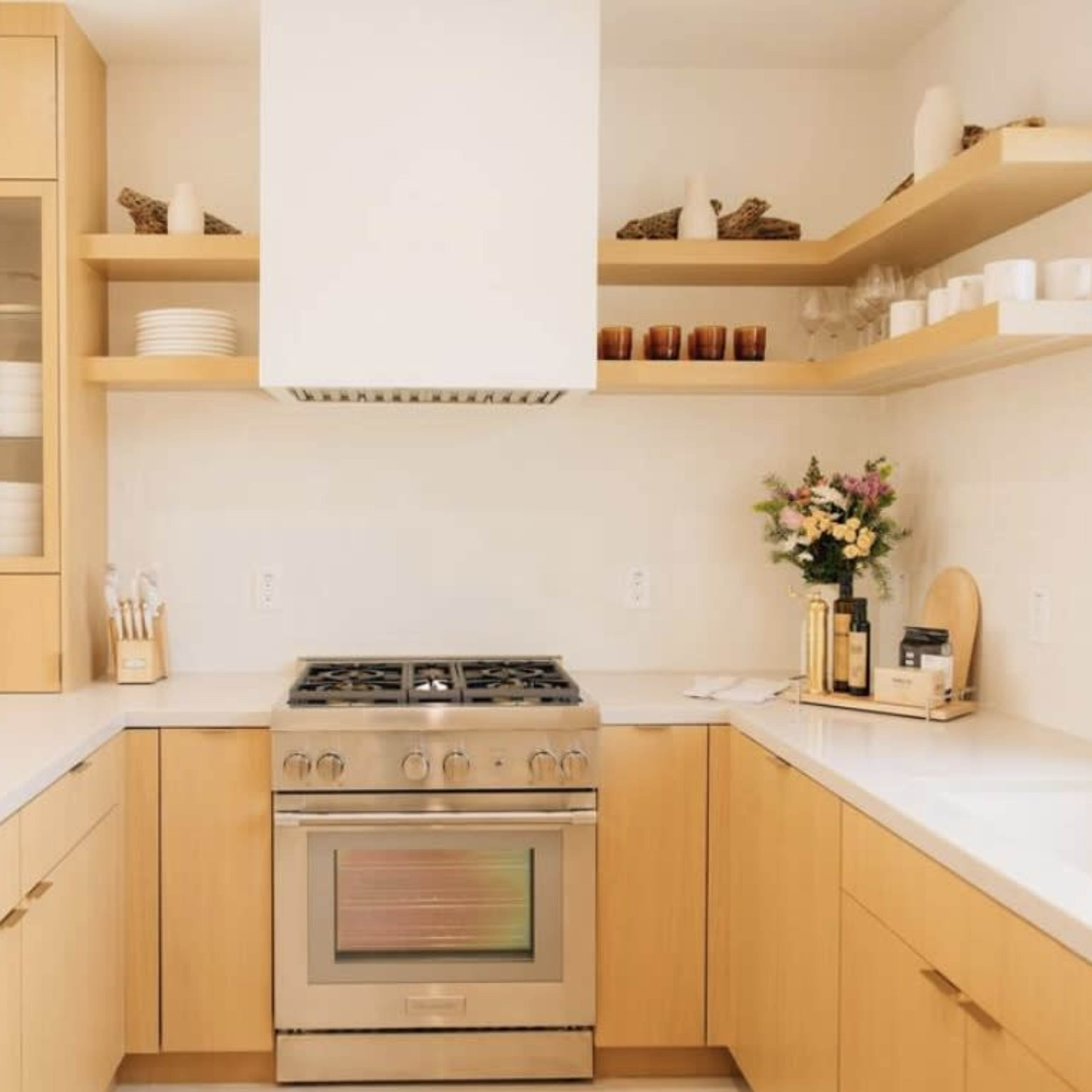A modern kitchen features light wood cabinets, a stainless steel stove, and open shelves displaying dishes and decorative items.