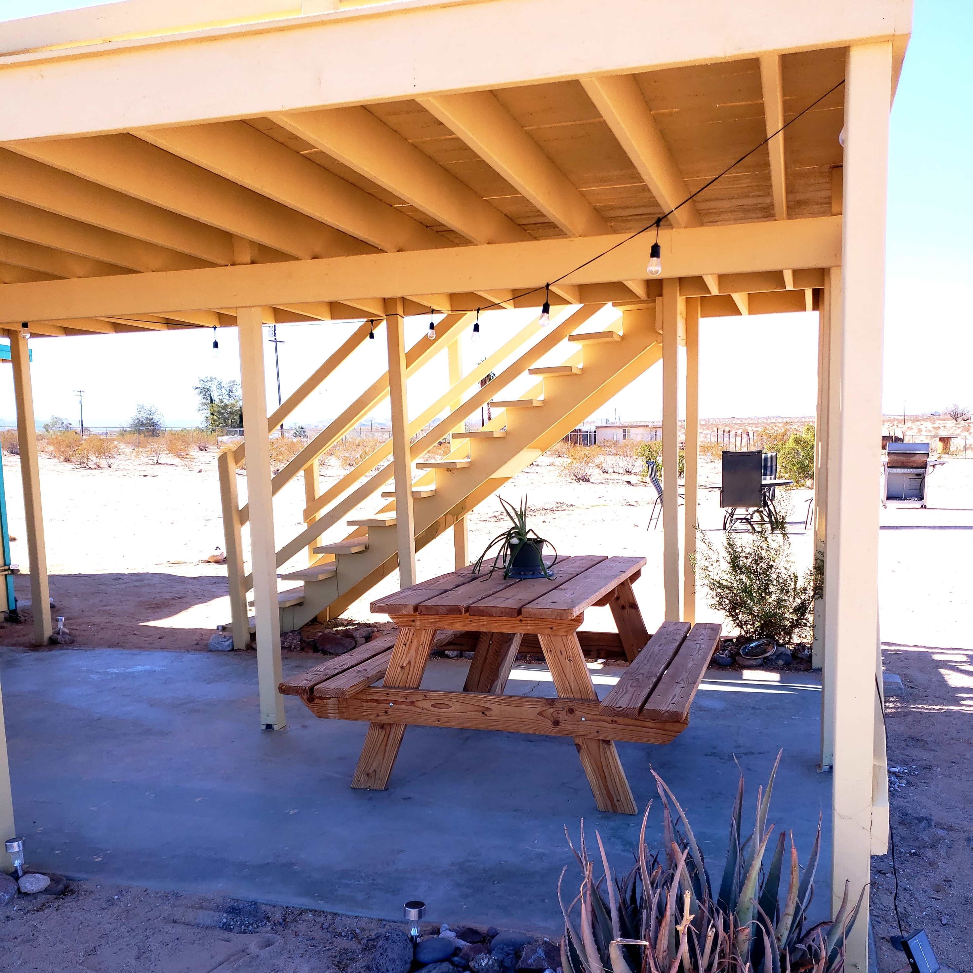 A wooden picnic table is situated beneath a yellow wooden structure with a staircase, surrounded by a desert landscape.