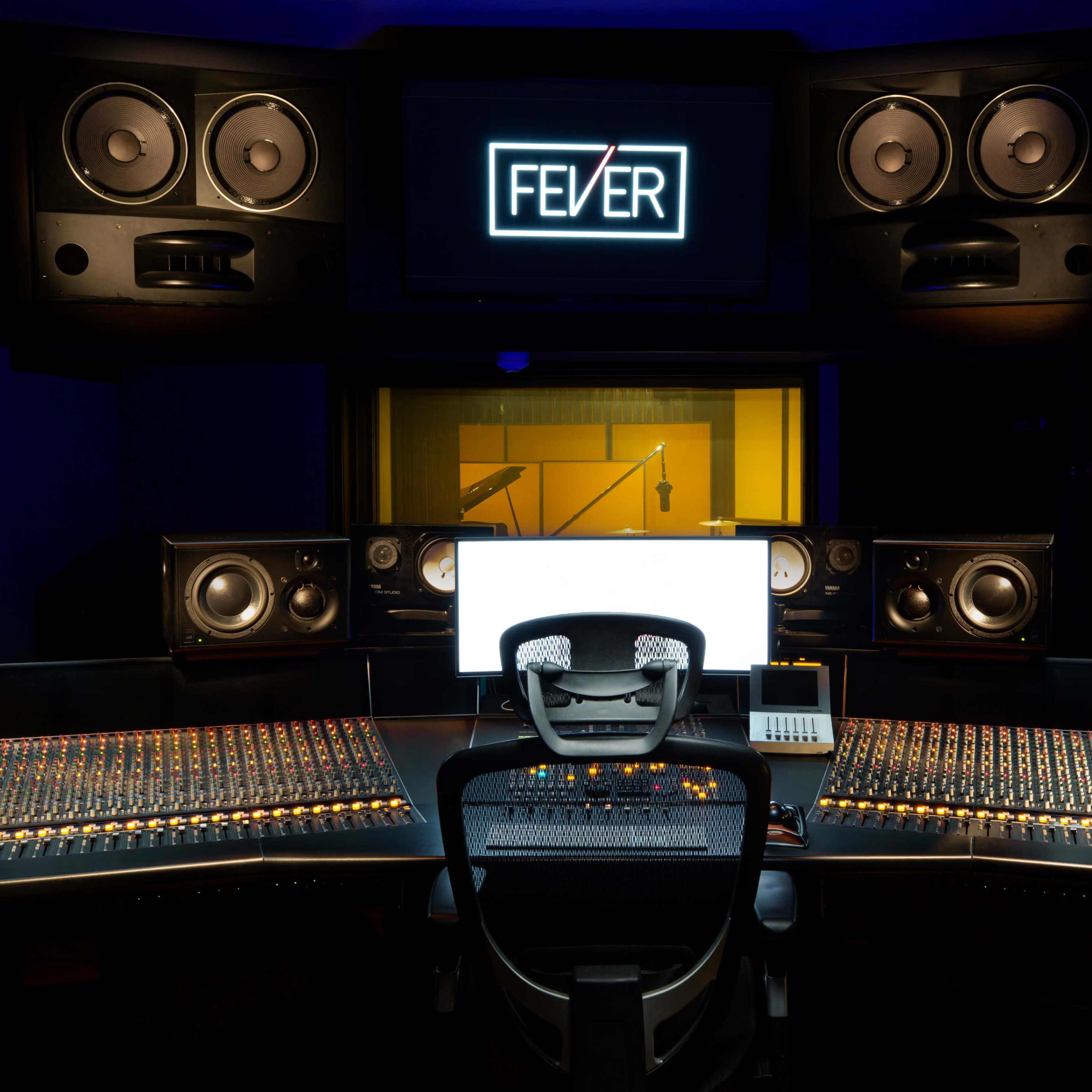 A professional audio recording studio features a large mixing console, multiple speakers, and a monitor displaying the word "FEVER."