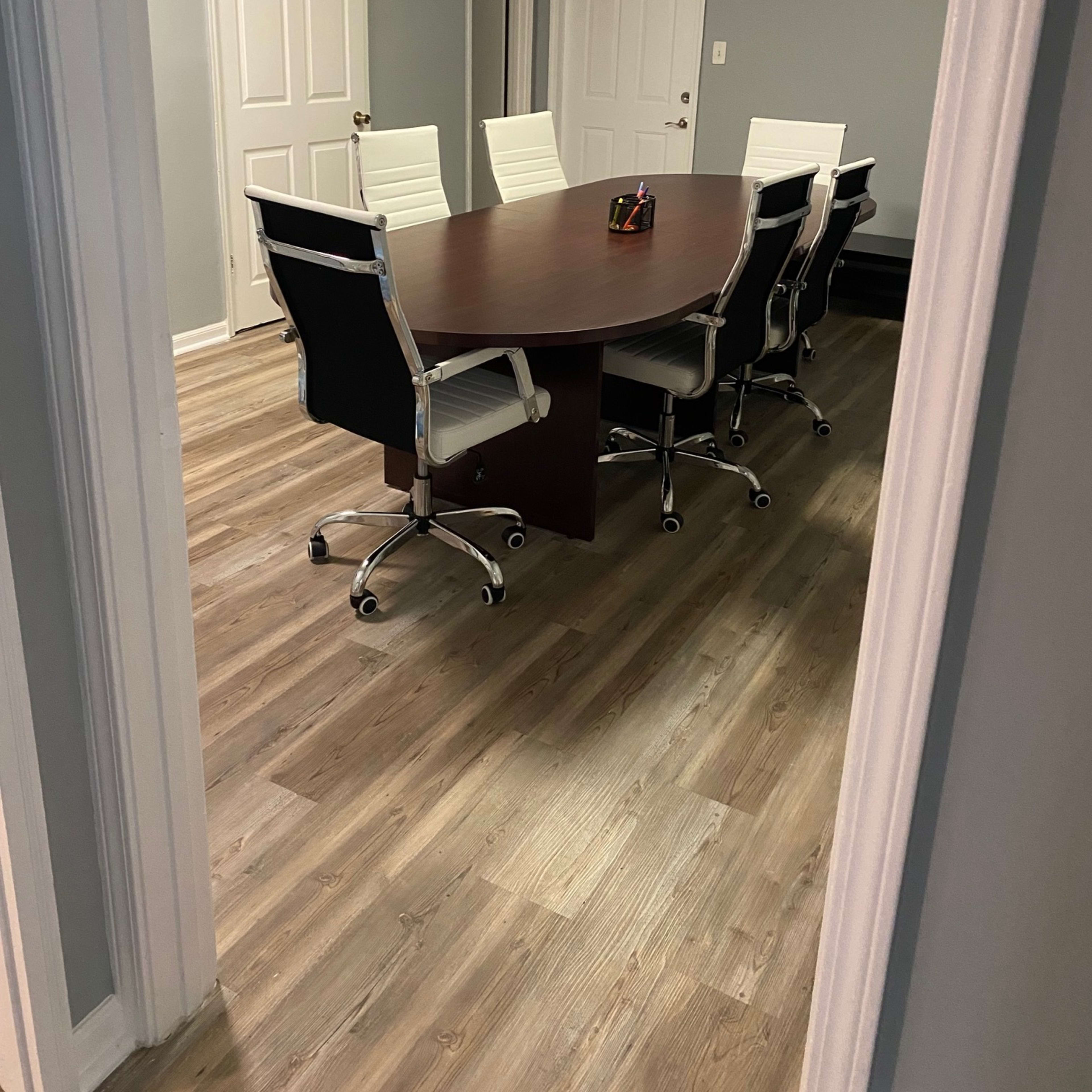 A conference room features a large oval table surrounded by white chairs on a wooden floor, with gray walls and two closed doors in the background.