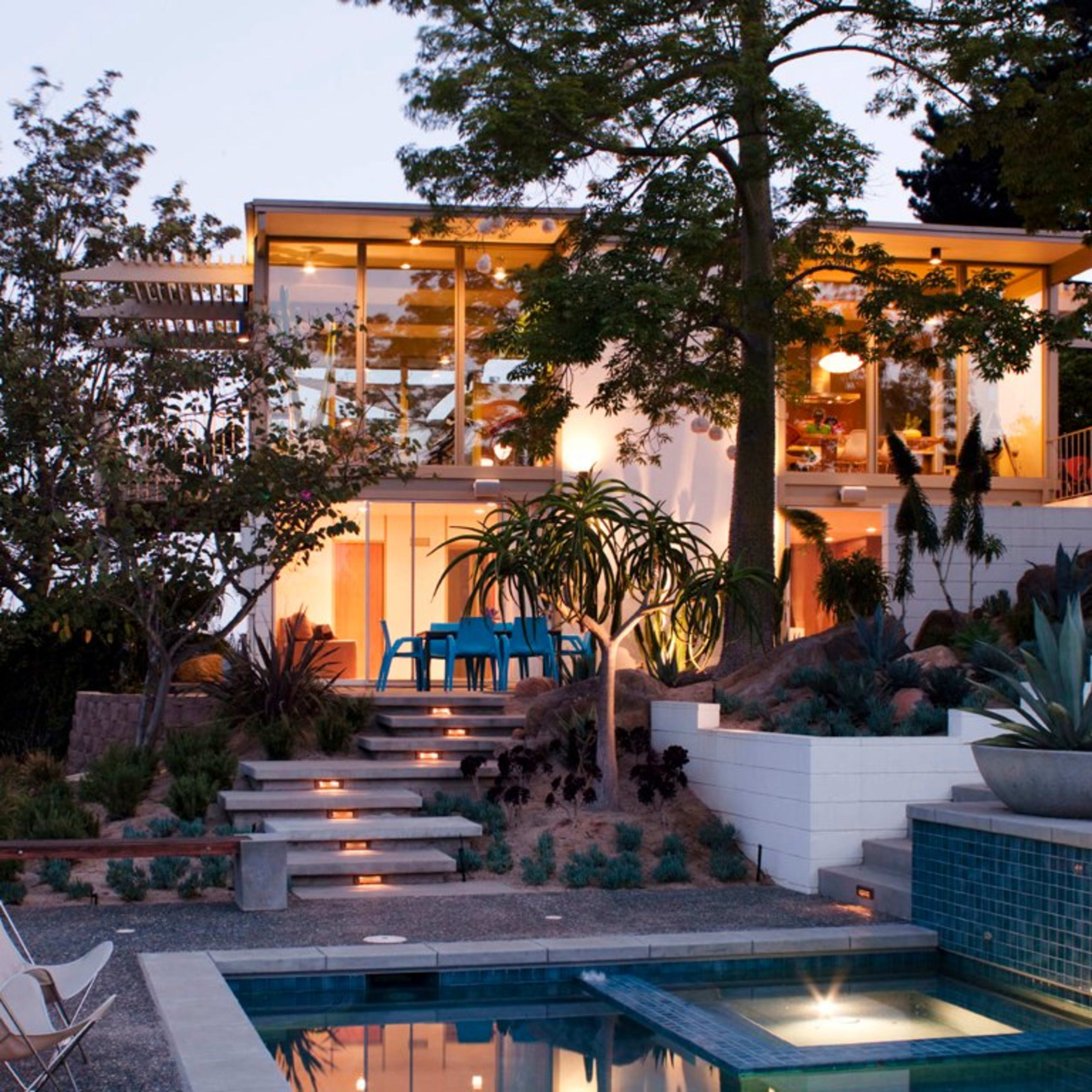 A modern house with large glass windows and a pool is surrounded by landscaped gardens and walkways leading up to it.