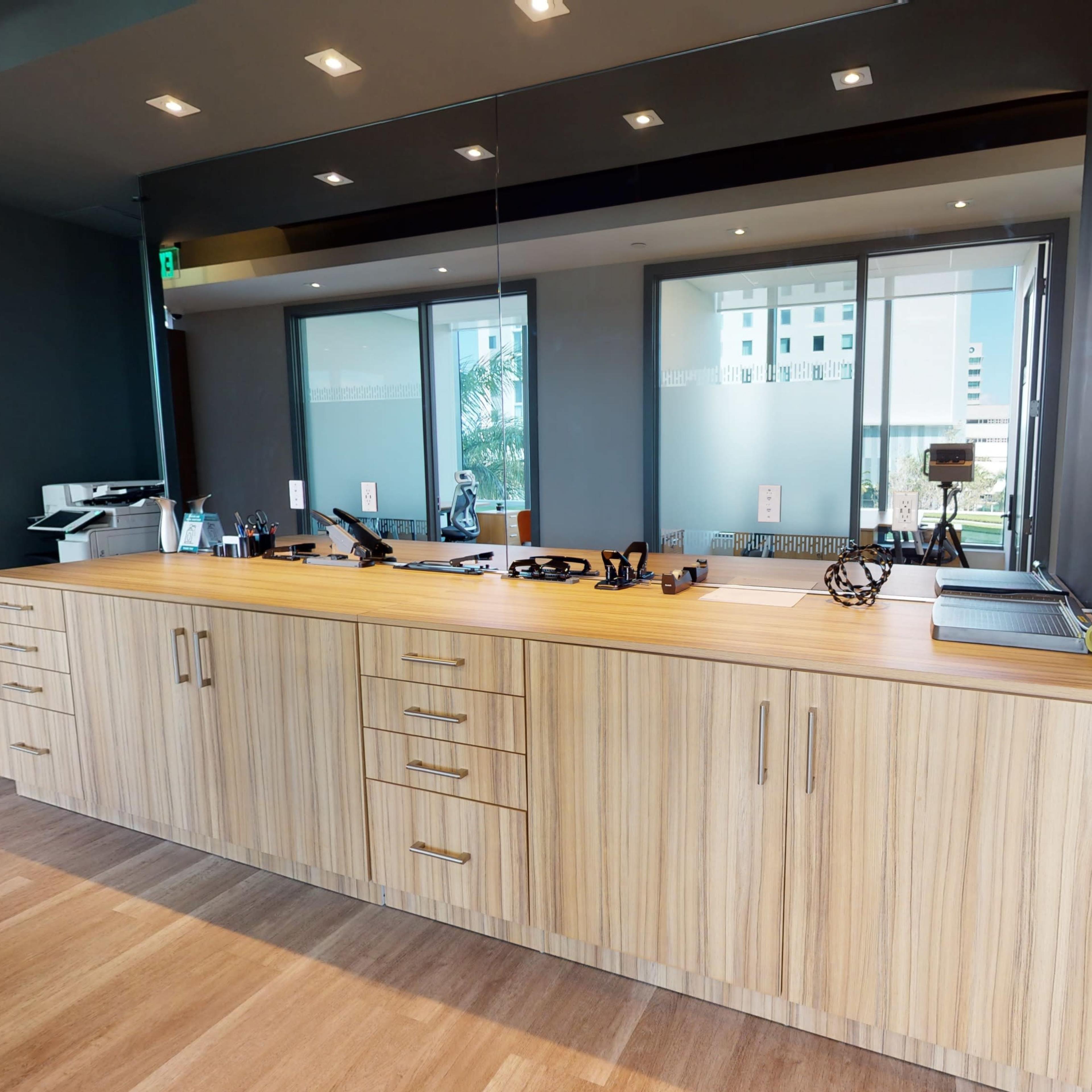 A modern reception desk with a wooden surface, equipped with various office supplies and a mirror backdrop, situated in an office space.