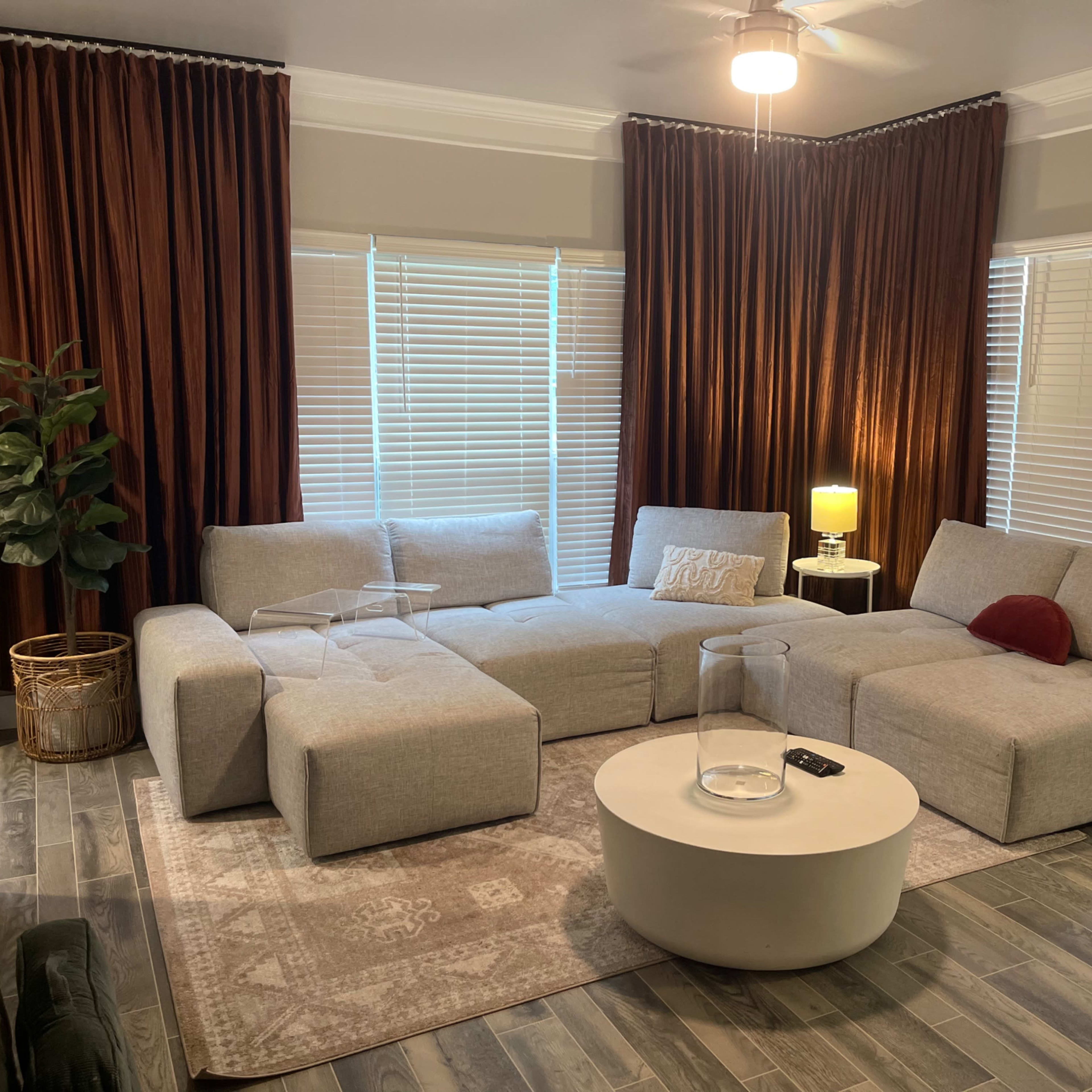 A modern living room featuring a large gray sectional sofa, a round white coffee table, and dark curtains, with a floor lamp beside the window.