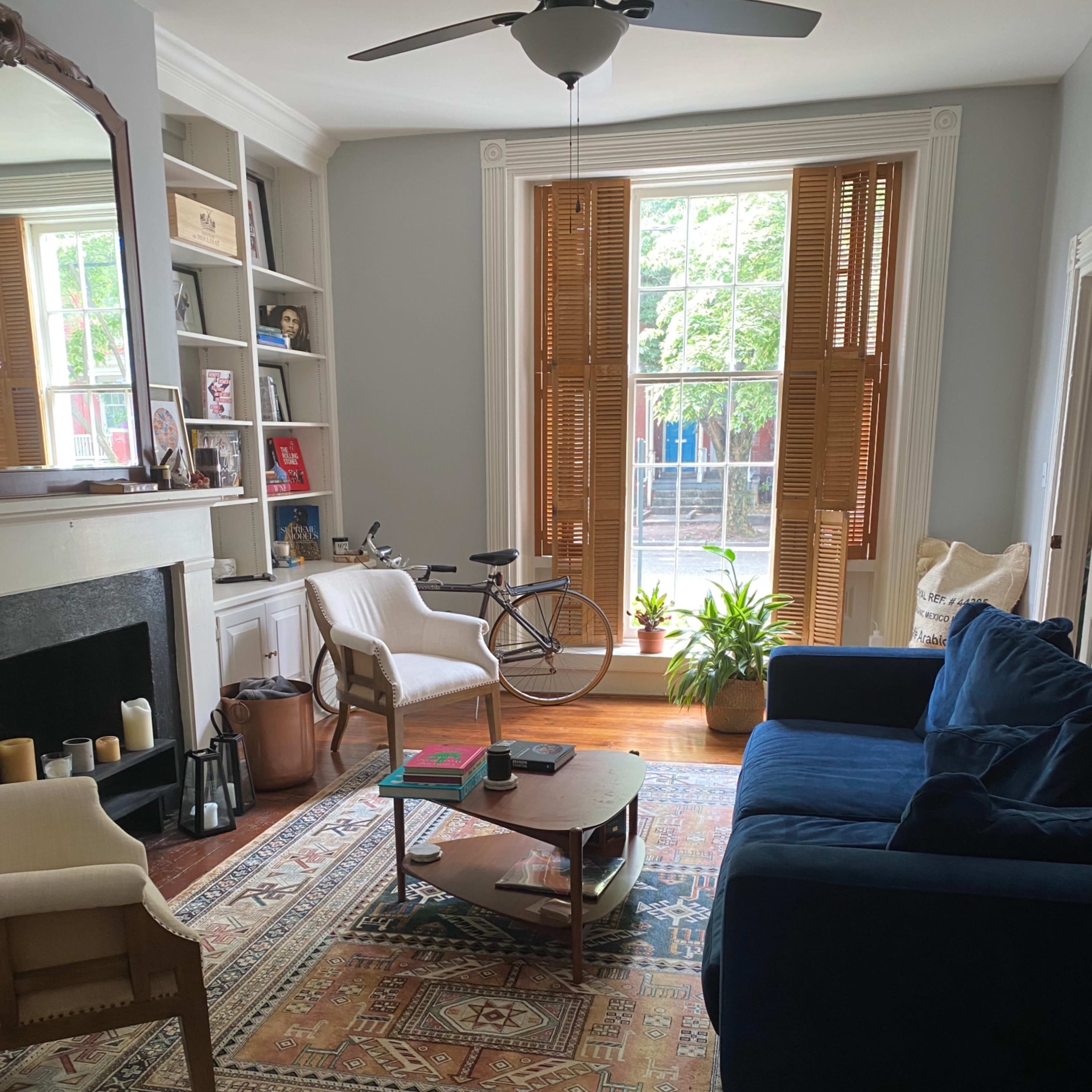 The living room features a blue sofa, a coffee table, a bicycle, and large windows with wooden shutters, all arranged around a patterned area rug.