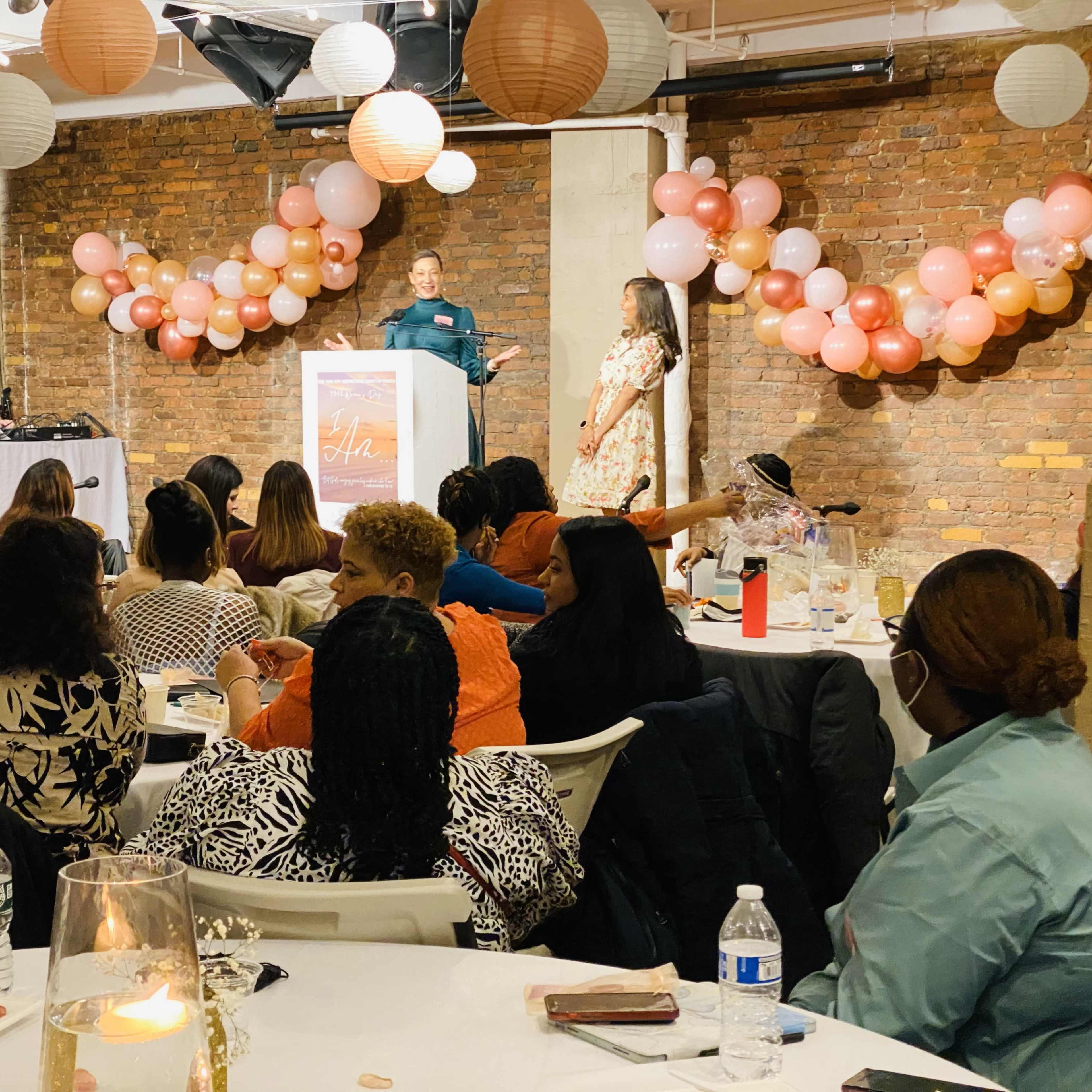 A speaker addresses an audience seated at tables decorated with balloons in a well-lit event space.