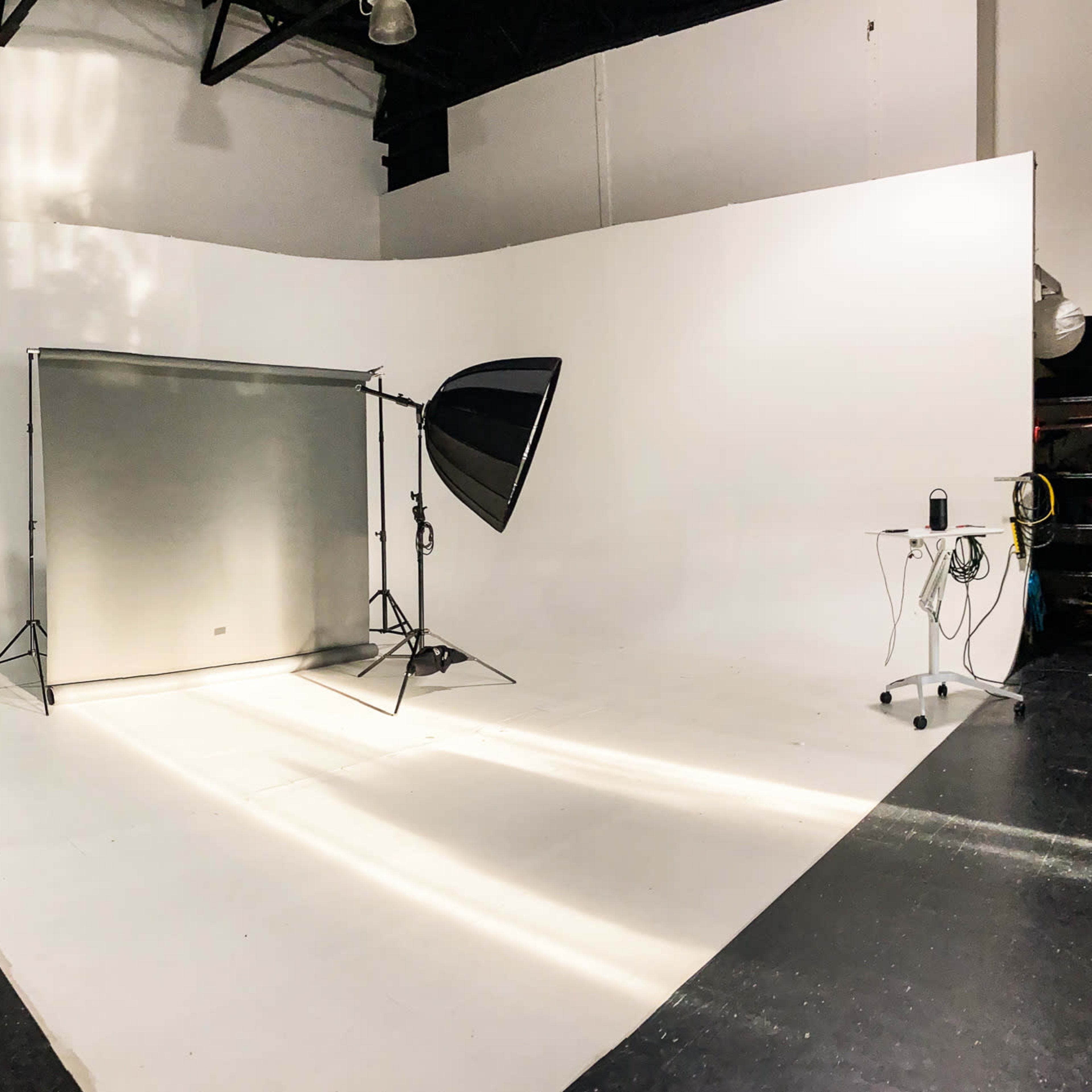A photography studio with a large, white backdrop, a light source on a stand, and a small table with equipment.