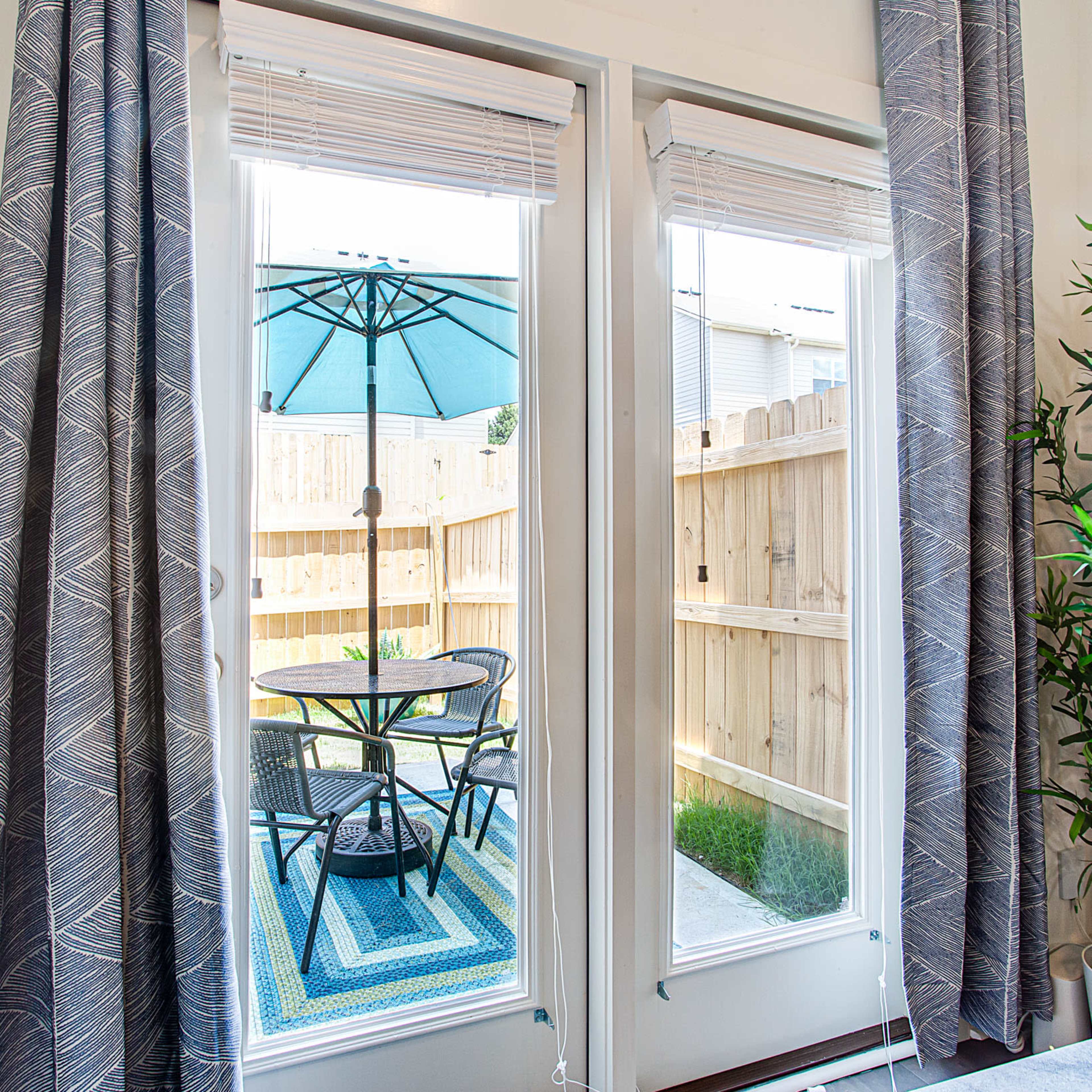 A patio with a table and chairs under an umbrella is visible through two glass doors, framed by patterned curtains.