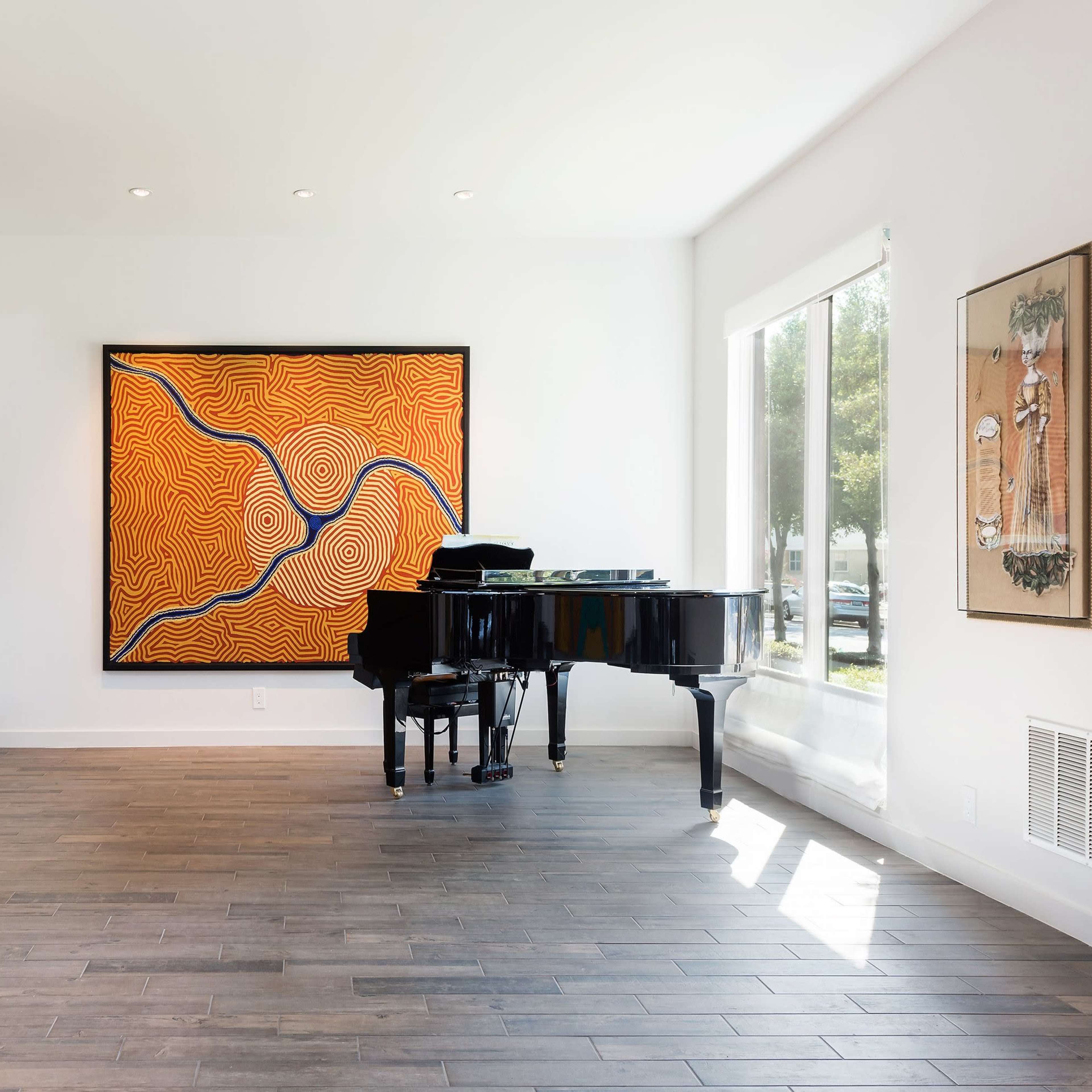 A black grand piano is positioned beside large windows in a room featuring colorful artwork on the walls and hardwood flooring.