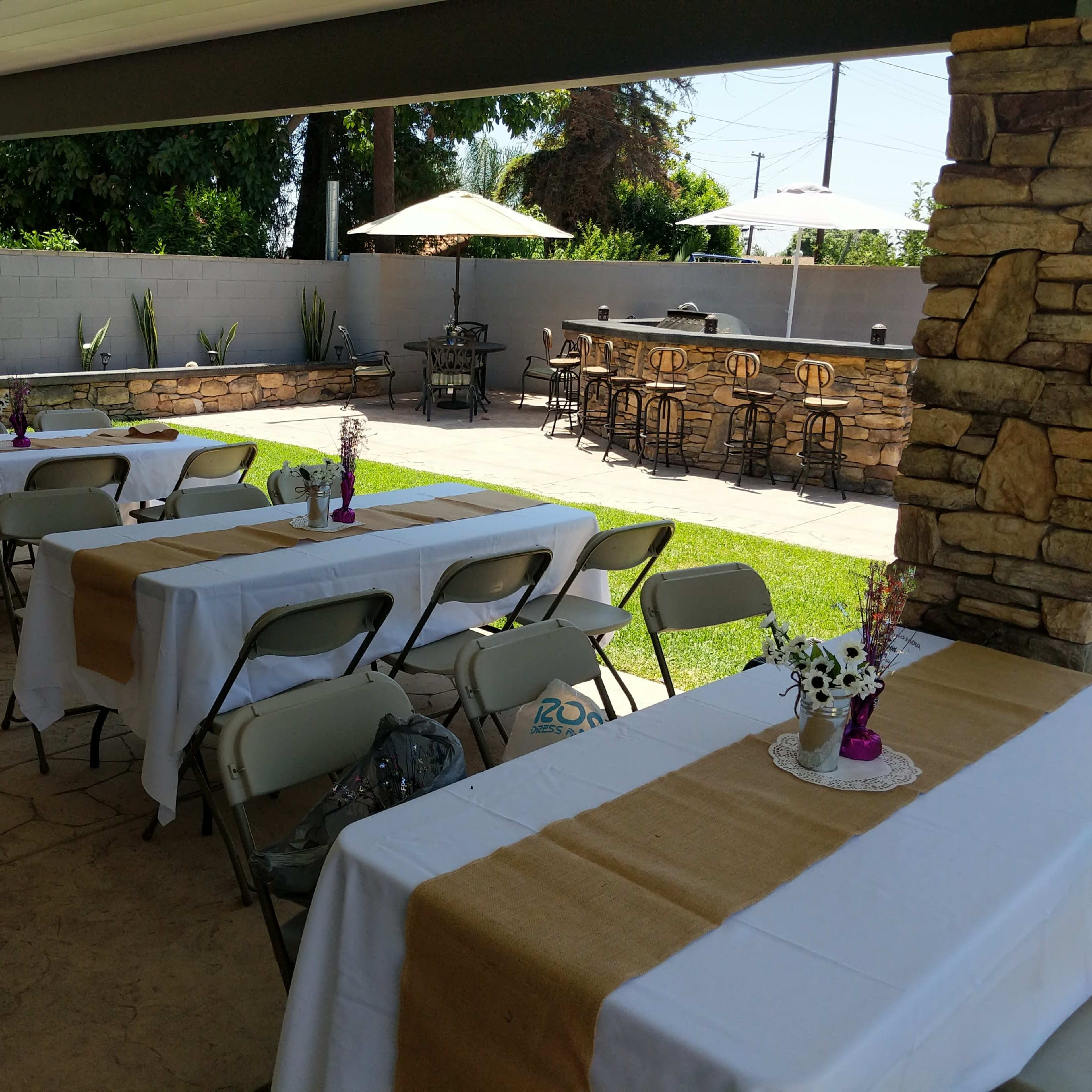 A shaded outdoor space features several tables covered with white tablecloths and brown runners, surrounded by stone columns and greenery, with a bar area in the background.