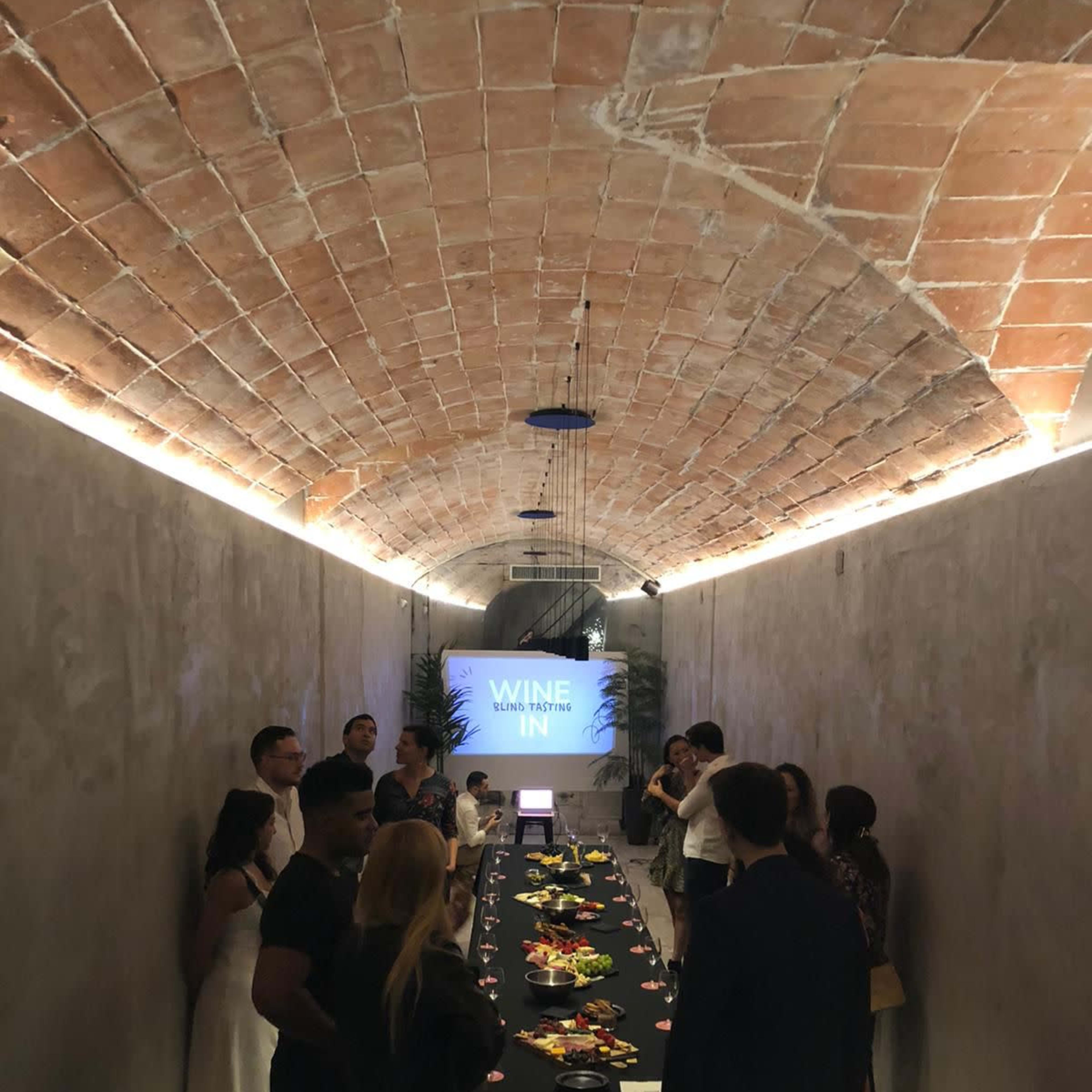 A group of people stands around a long table laden with food in a well-lit, arched room displaying a "WINE IN" sign.