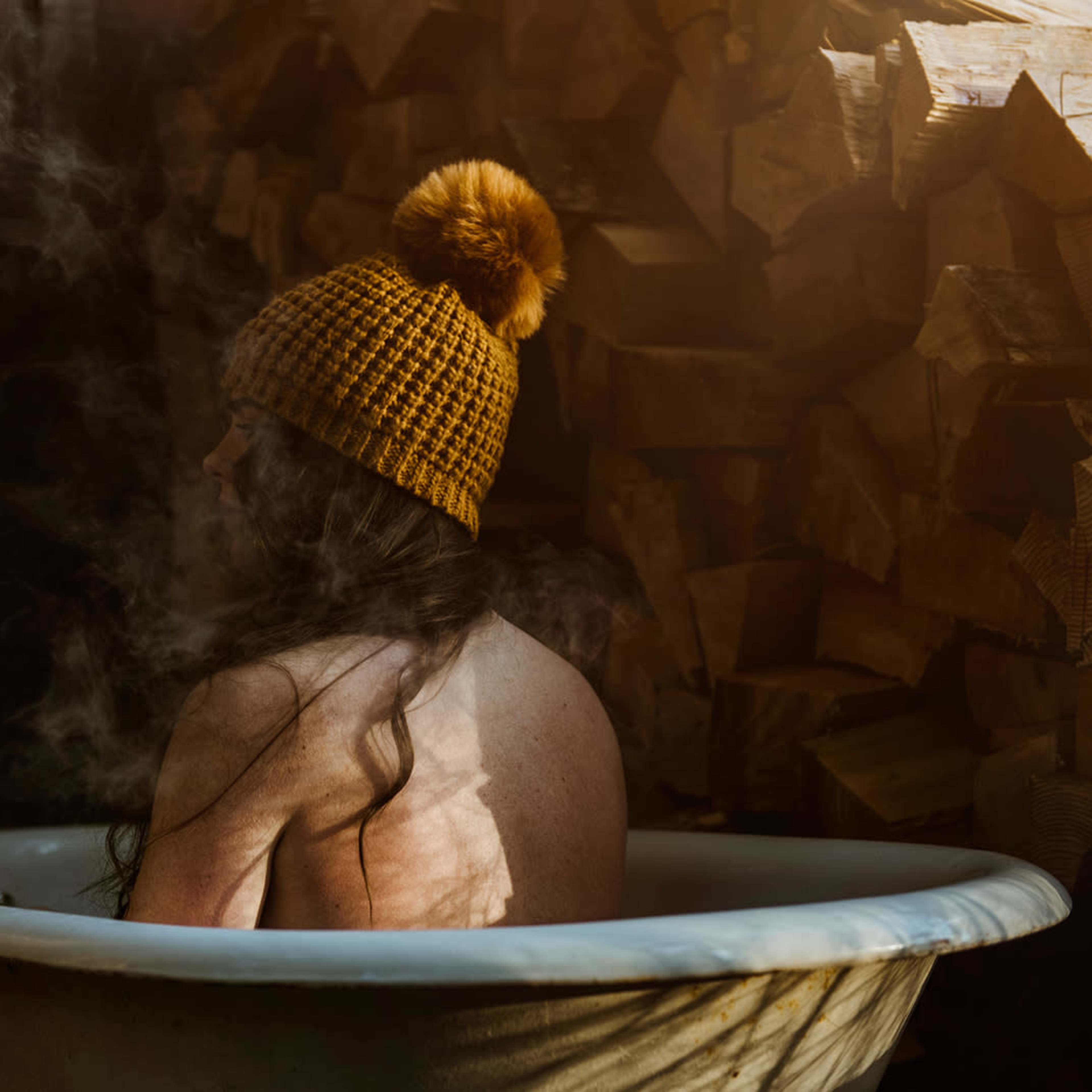 A person with long hair sits in a vintage bathtub surrounded by stacked firewood, steam rising around them.
