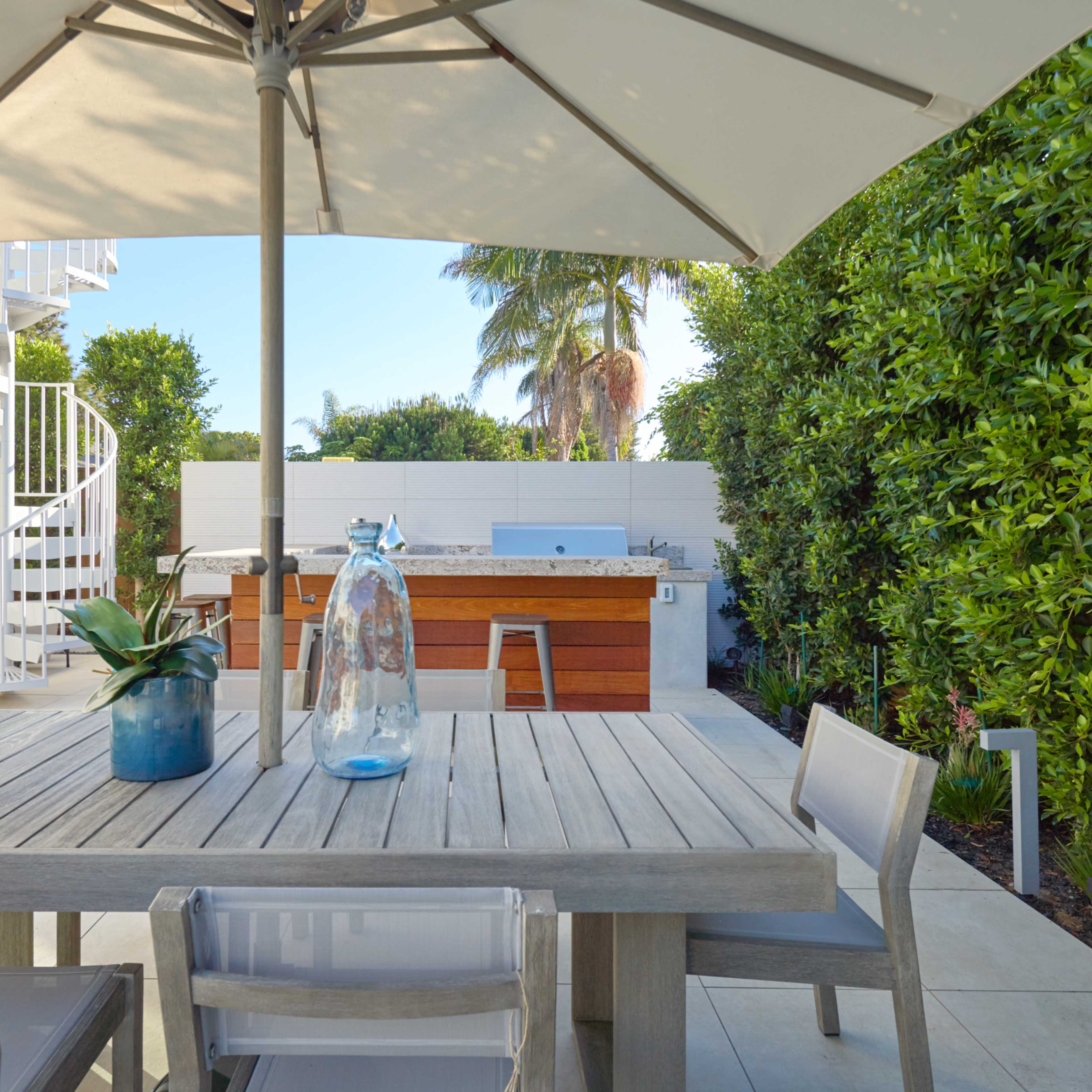 A wooden table with clear chairs is set under a large umbrella, with a potted plant and water jug on the table, overlooking an outdoor kitchen surrounded by greenery.