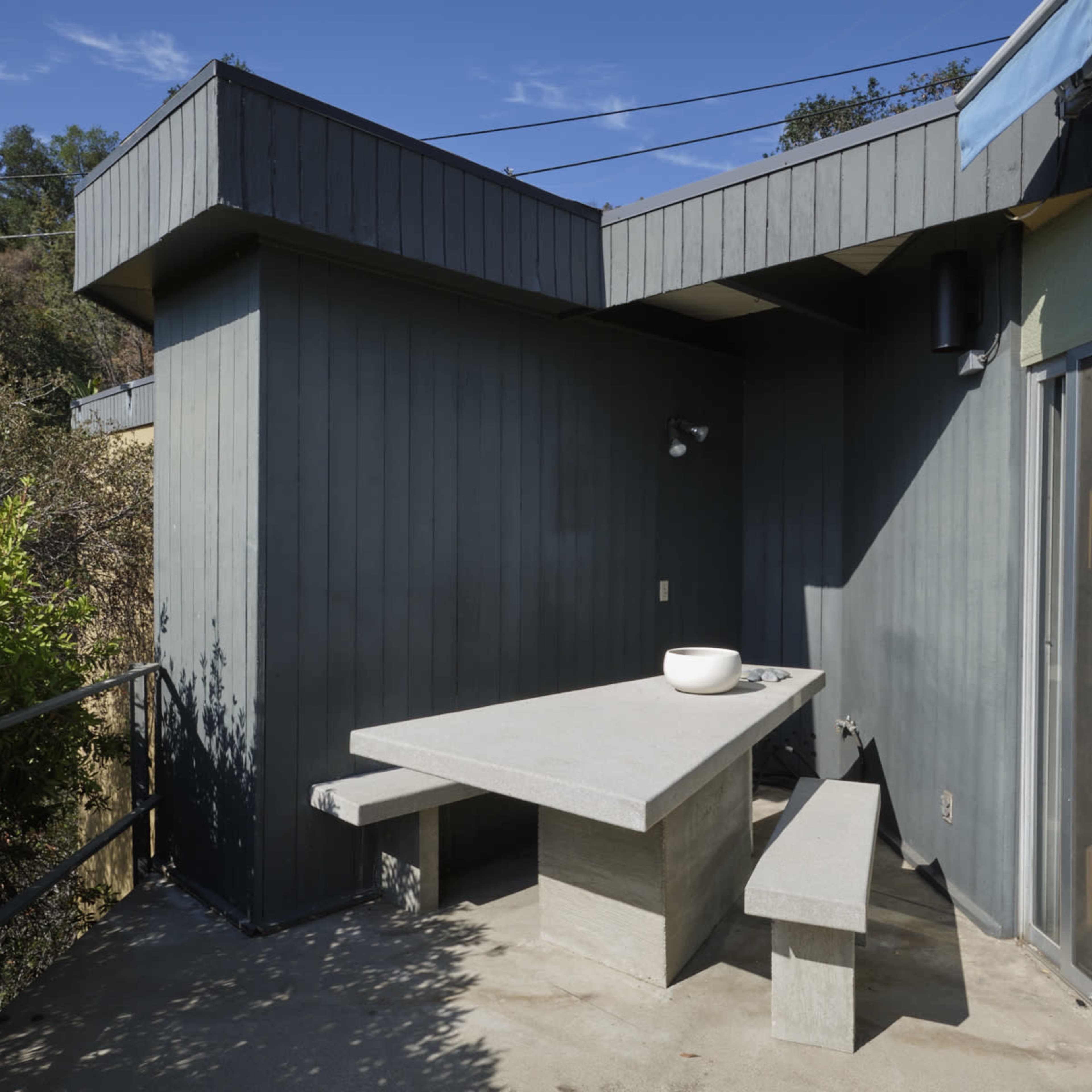A concrete table and bench on a patio surrounded by greenery, with a building featuring a slanted roof in the background.