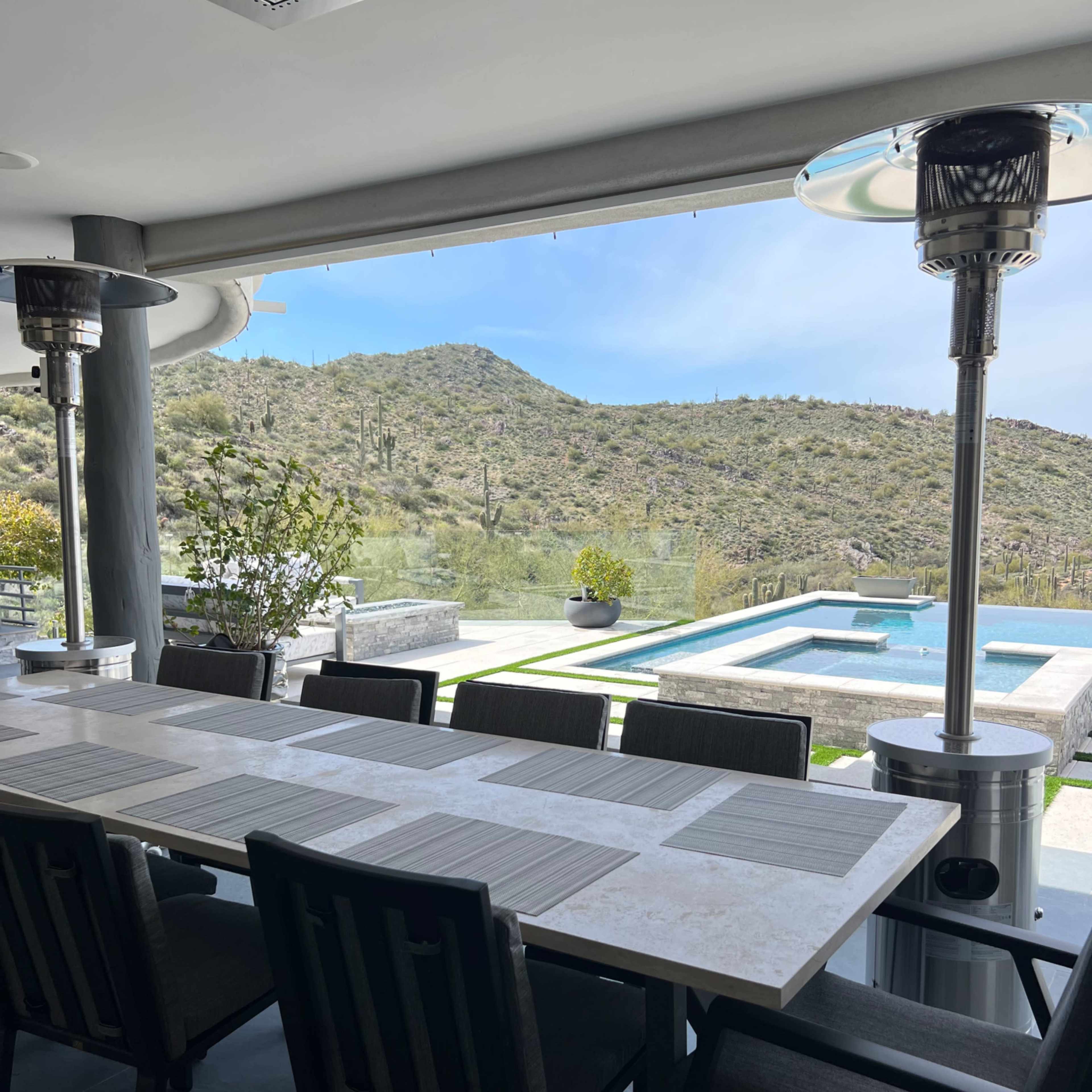 A dining table is set on a patio overlooking a pool and mountainous desert landscape.