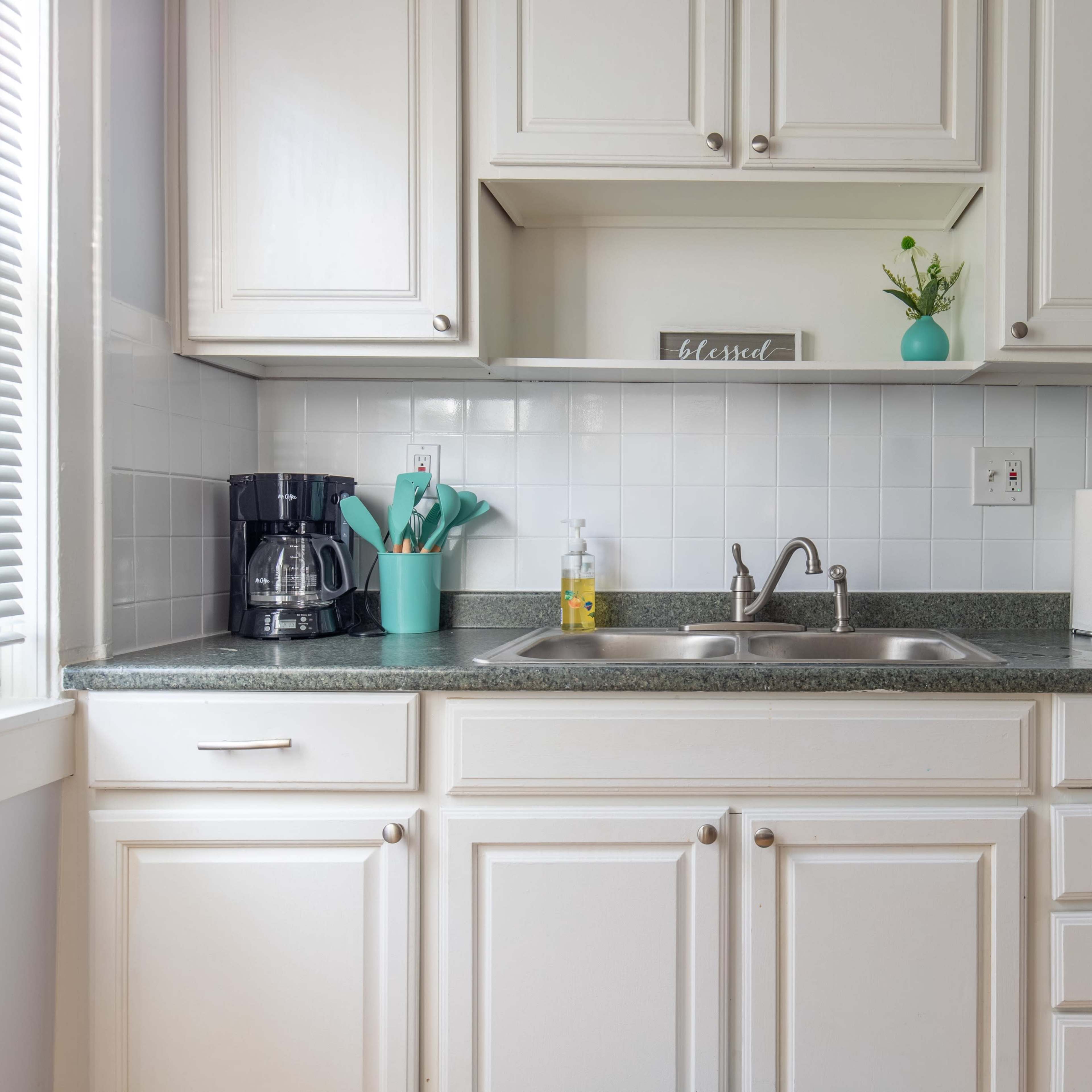 A clean kitchen counter features a coffee maker, a stainless steel refrigerator, a sink with dish soap, and neatly organized utensils in a turquoise holder.