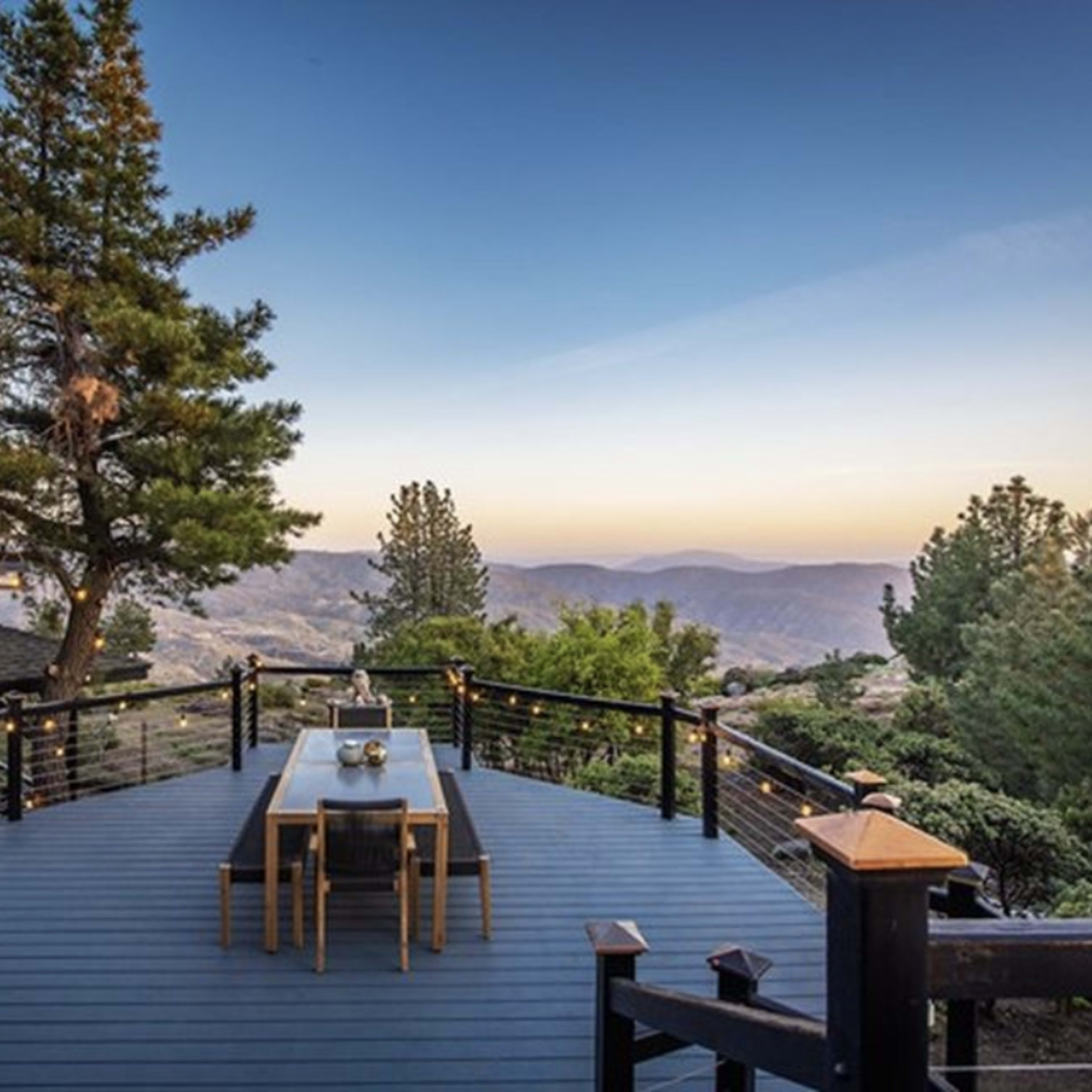 A wooden deck with a long table overlooks a mountainous landscape at dusk, surrounded by trees.